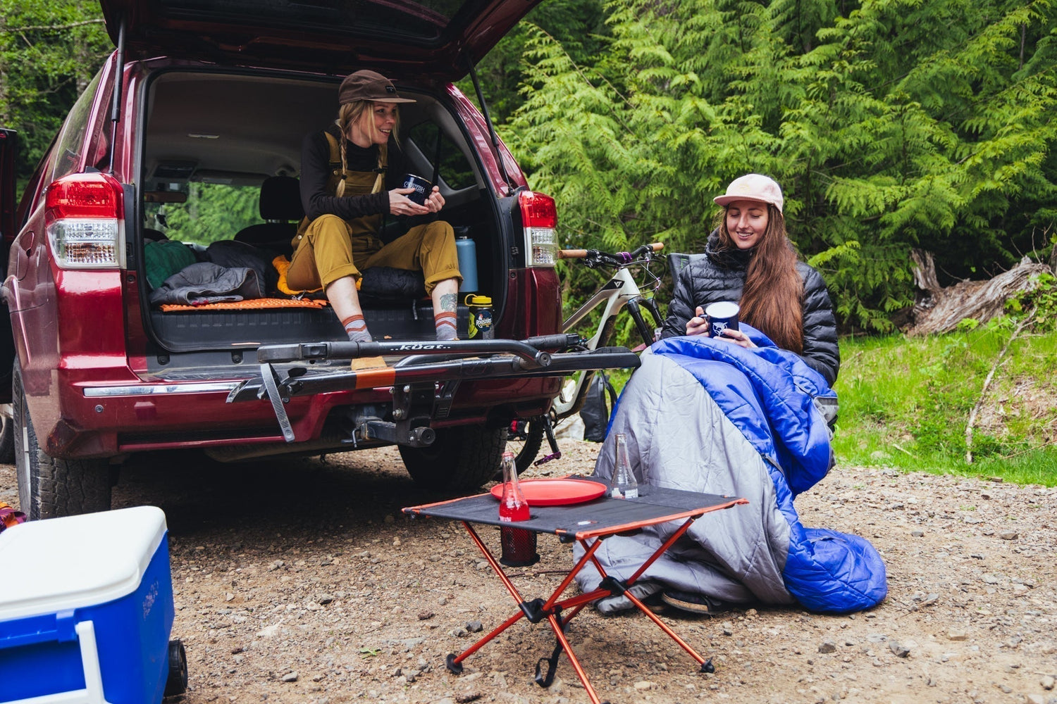 Two ladies setting up camp out of the trunk of a red car with cooler, chairs and mini table set up in dirt