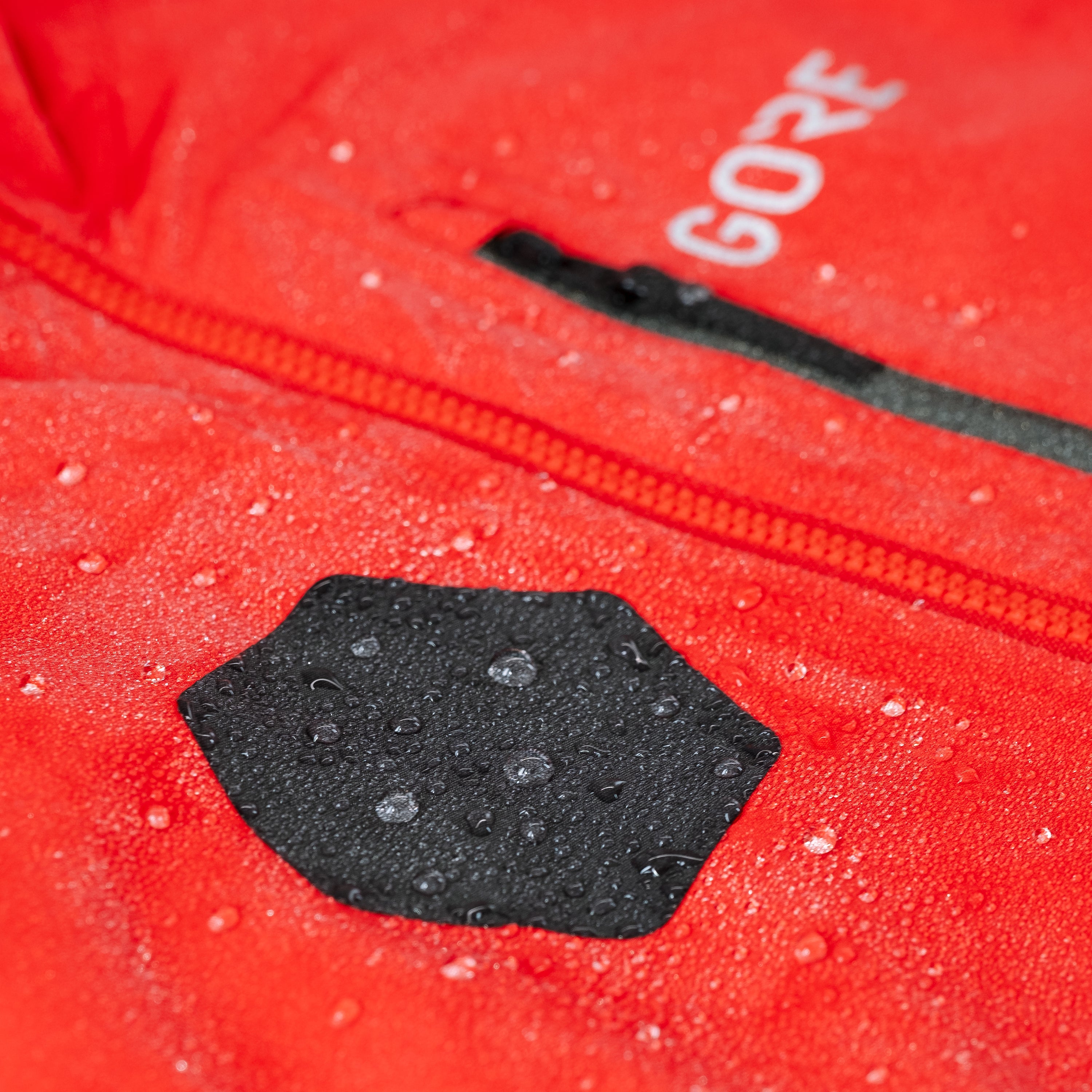Red jacket with a black patch and 'GORE' branding, showing water droplets.