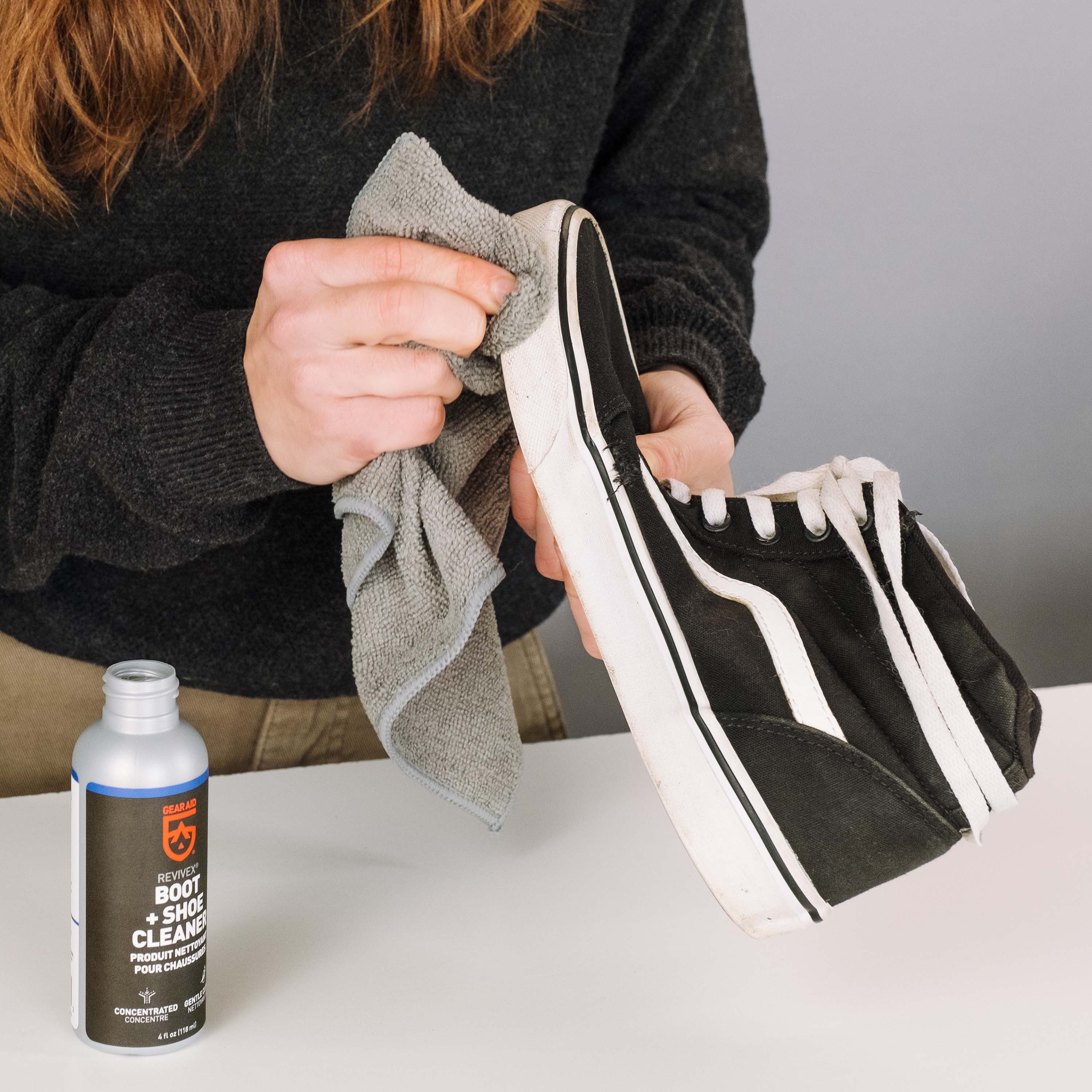 Person cleaning a black and white sneaker with a cloth, next to a bottle of boot and shoe cleaner.