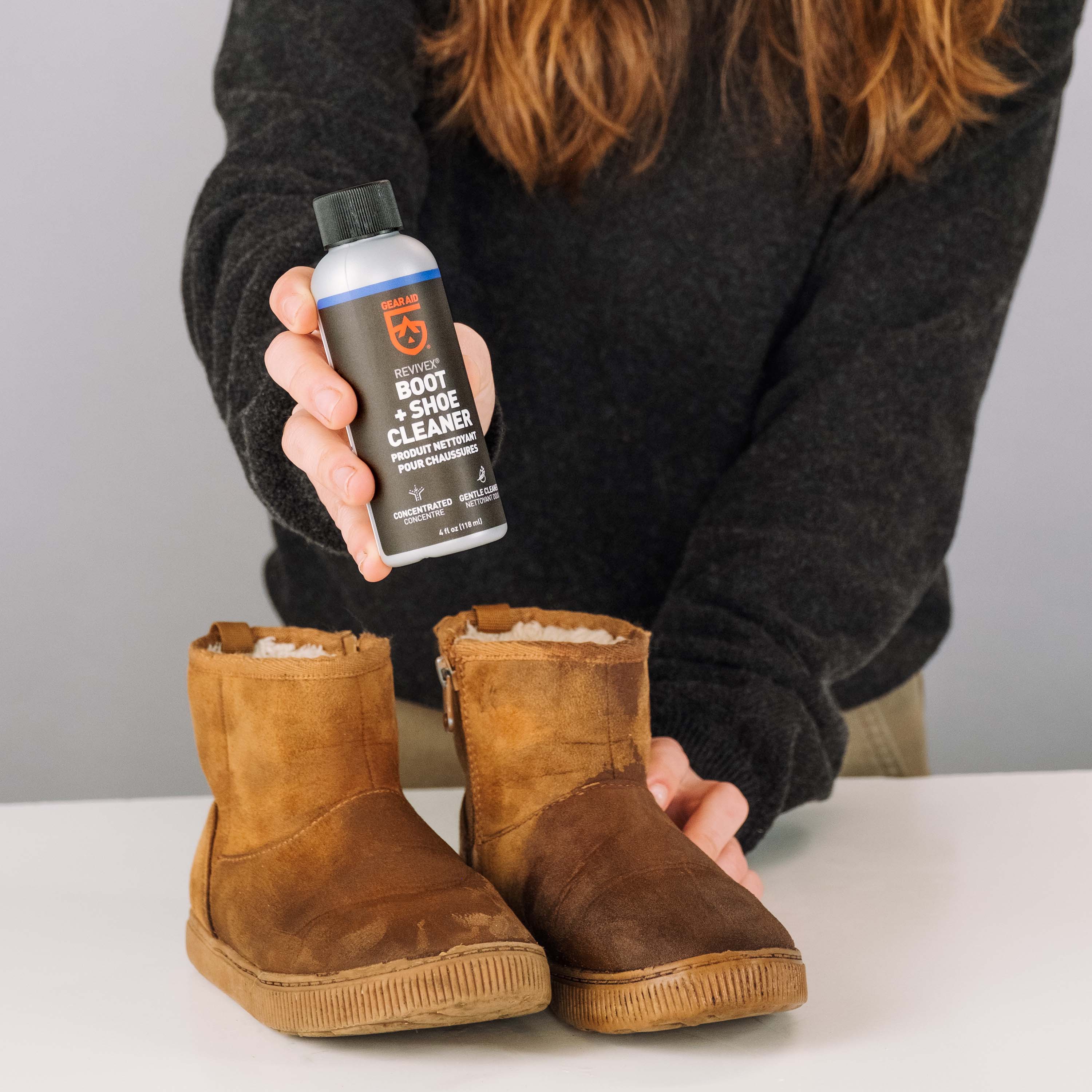 Person holding a bottle of boot cleaner next to worn brown boots on a white surface.