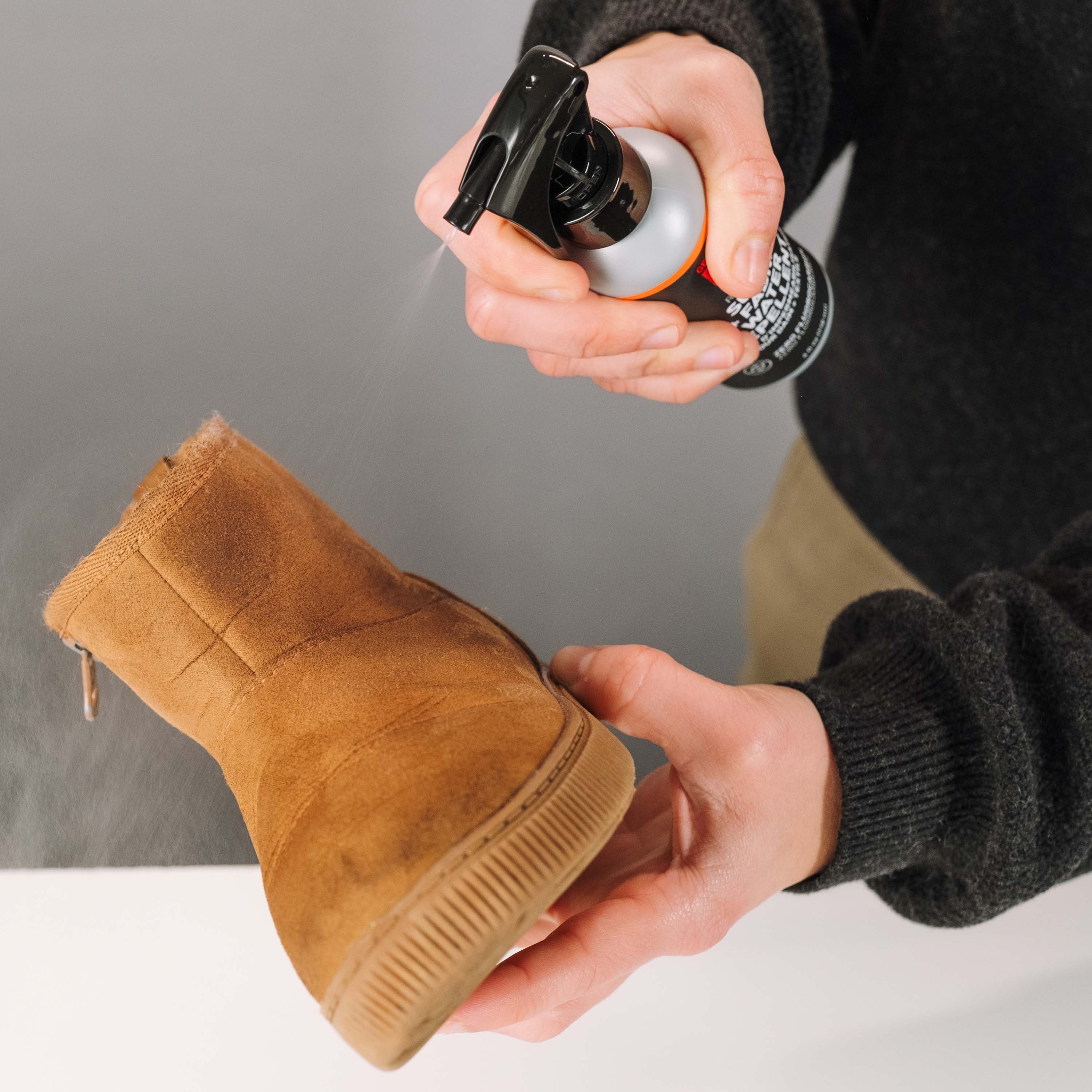 Person cleaning a brown suede boot with a spray bottle against a neutral background