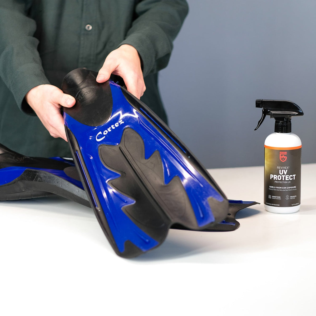 Person holding a blue and black diving fin with a bottle of UV Protect spray on a white surface.