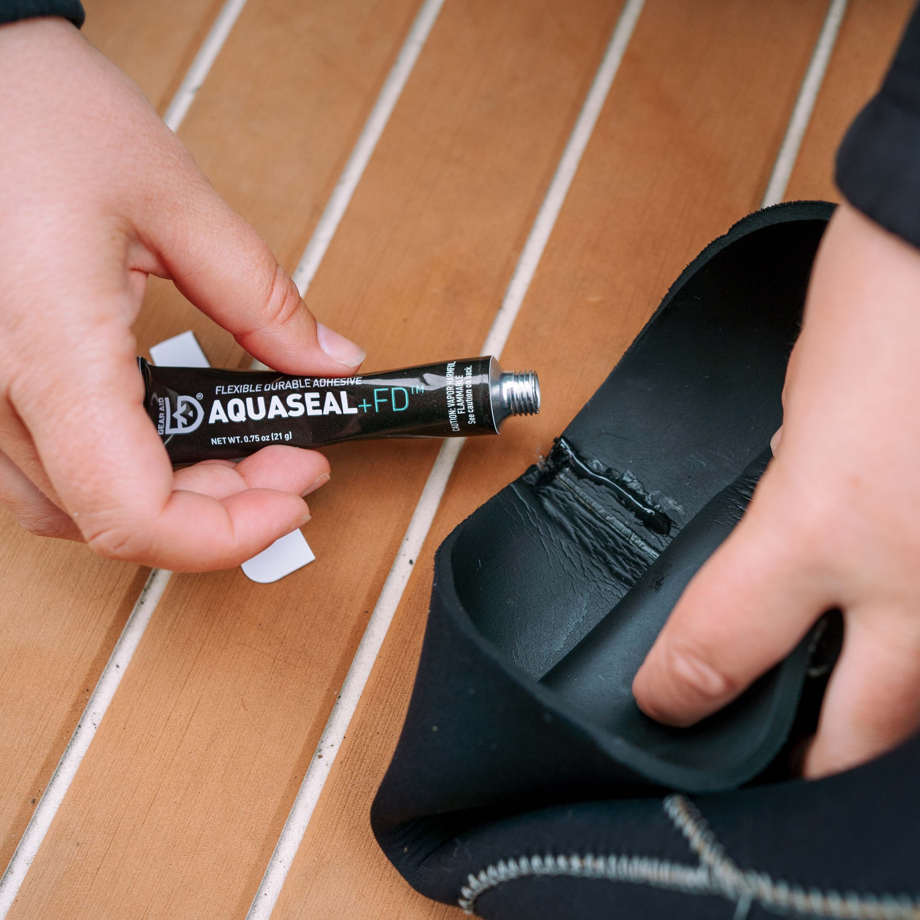 Person applying Aquaseal+FD to a black neoprene wetsuit neck gasket a wooden boat floor