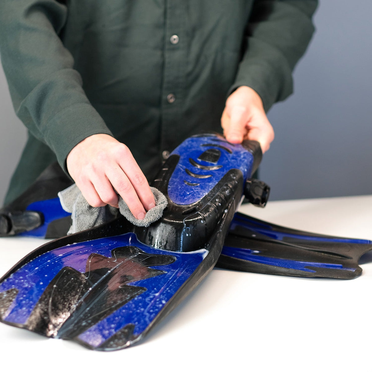 Person cleaning a pair of blue and black diving fins with a gray cloth on a white surface.