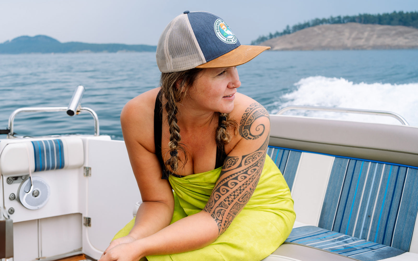 Woman with tattoos sitting on a boat wearing a green towel and cap with a logo, with water and mountains in the background.