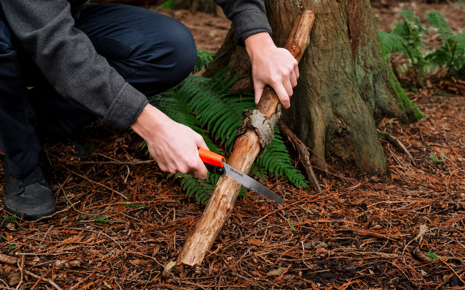 Person cutting a branch with a GEAR AID SKARA Camp Saw in a forest setting.