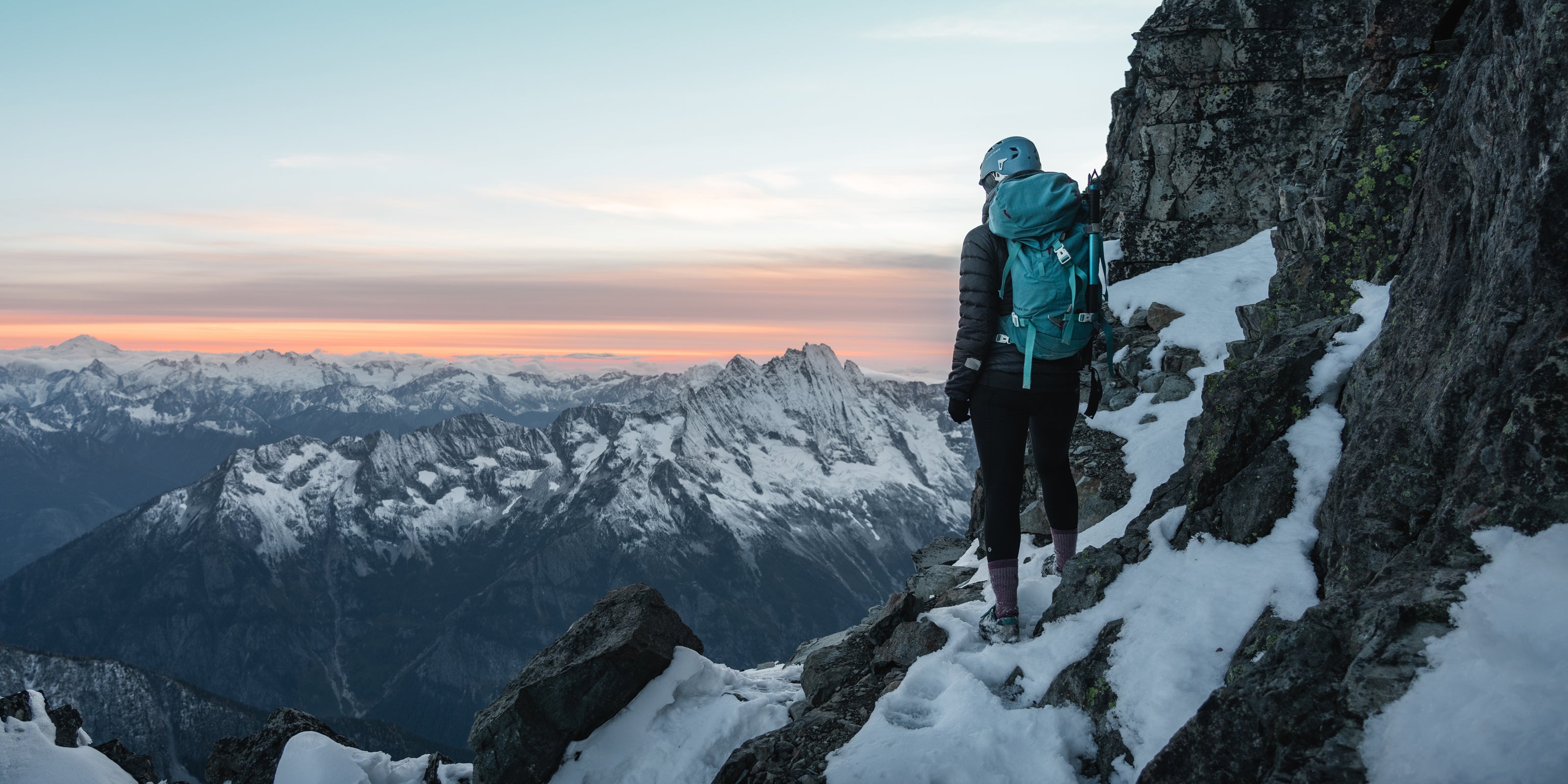 Person with a backpack standing on a snowy mountain peak at sunset.