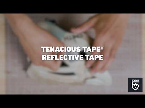Blurred video cover showing hands applying reflective tape to a light-colored bike helmet on a tabletop, with the title “TENACIOUS TAPE® REFLECTIVE TAPE” centered and the Gear Aid logo in the bottom-right corner.