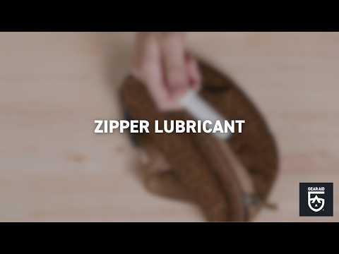 Blurred video cover showing a hand applying lubricant to the zipper of a brown pouch, with the title “ZIPPER LUBRICANT” centered and the Gear Aid logo in the bottom-right corner.