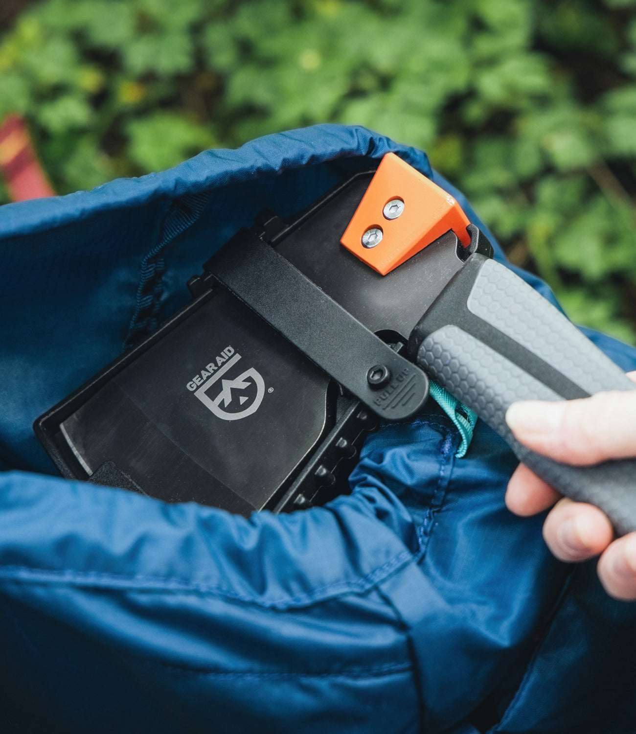 Person holding a camp hatchet with a Gear Aid logo on a blue backpack against a green shrub background