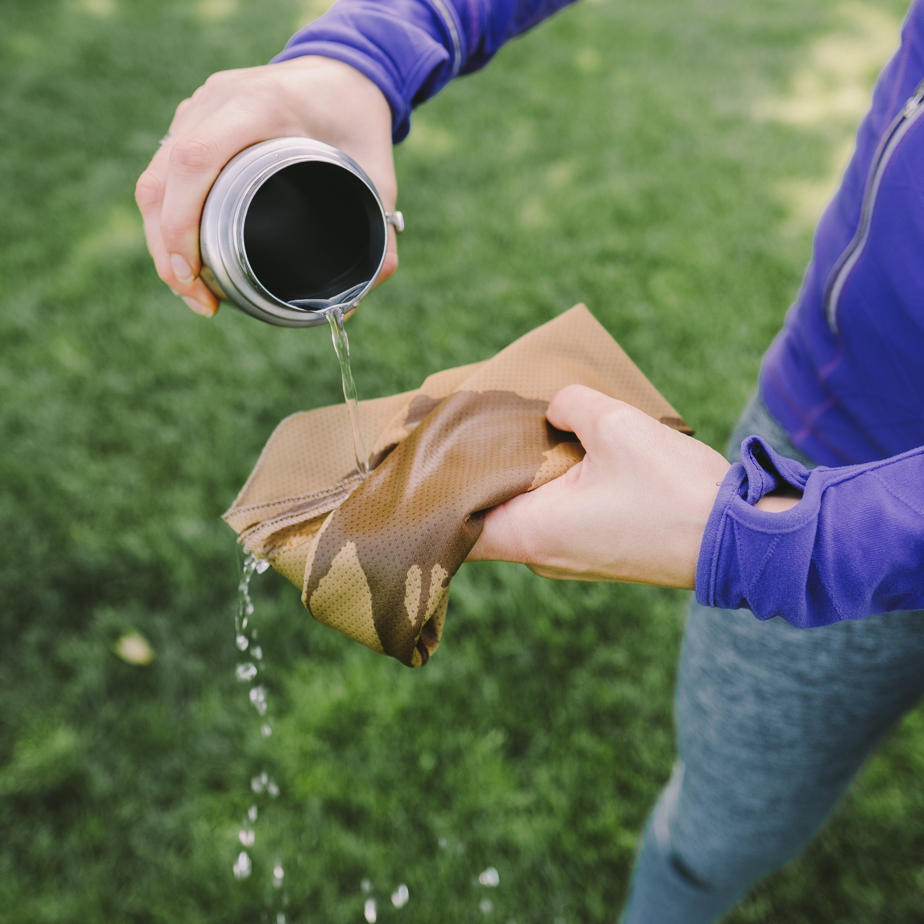 Person pouring water from a Hydroflask onto a cooling towel with grass in the background