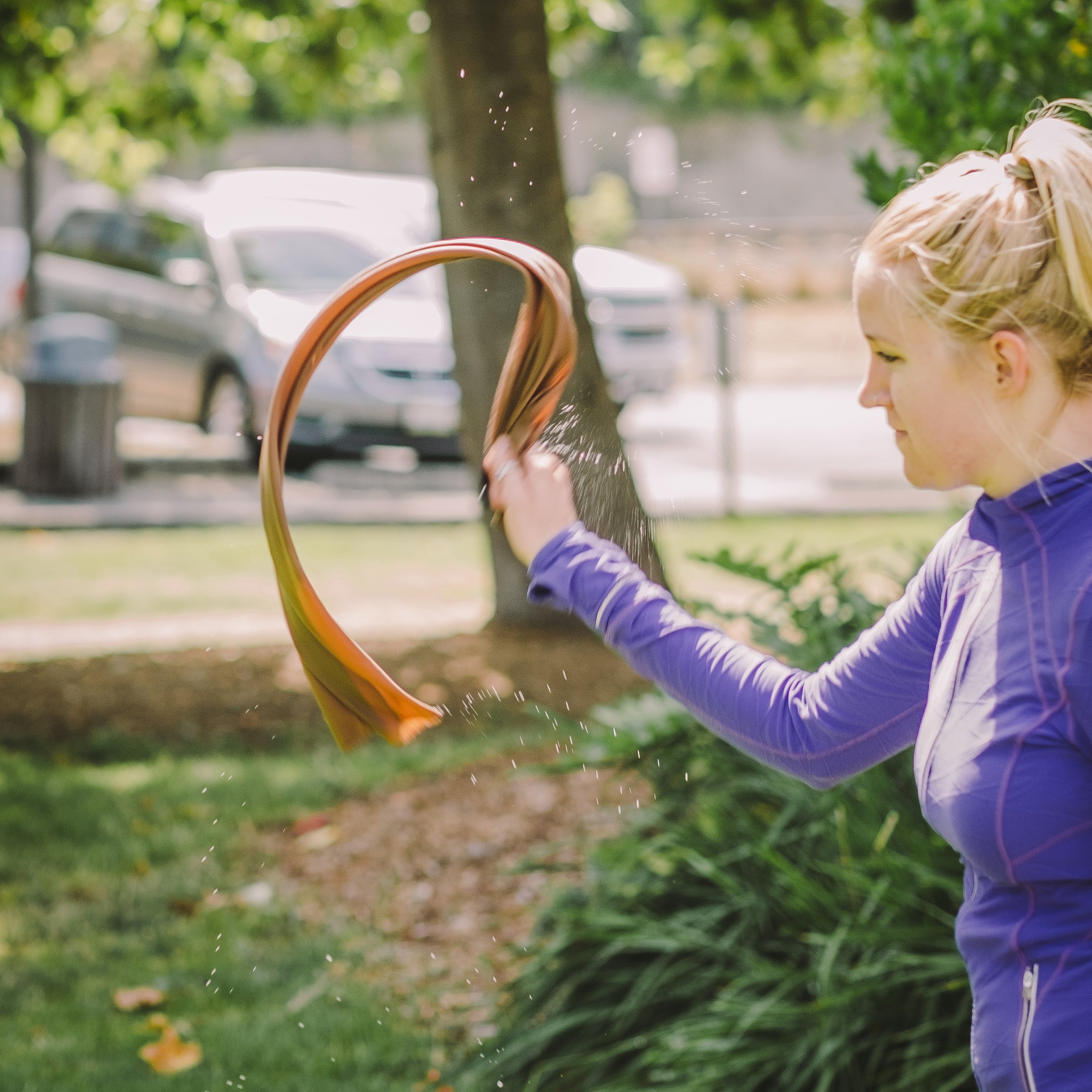 Person wearing purple and whipping/snapping a cooling towel in the air to activate it, trees and cars and bushes in the background