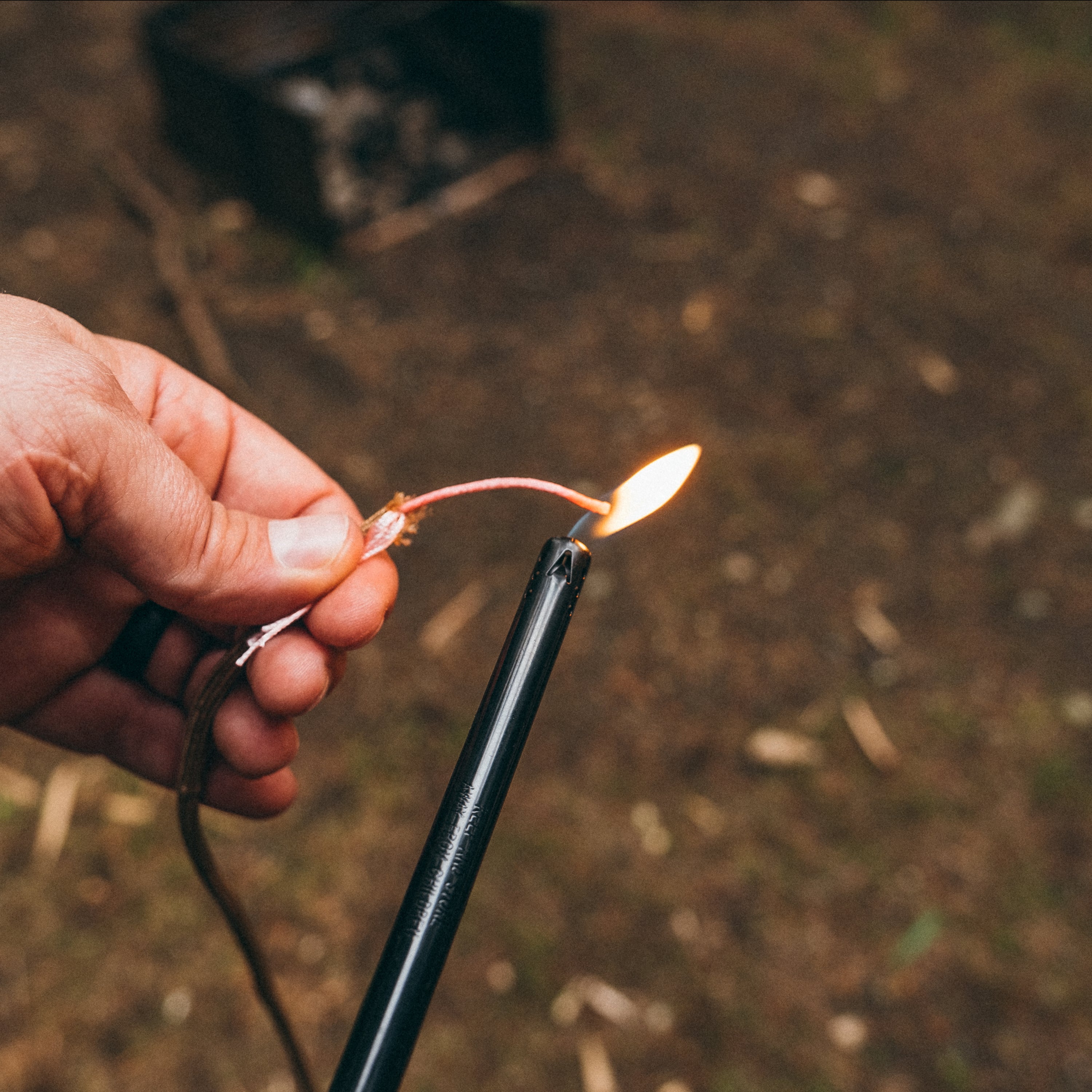 Paracord fire strand being lit up with a lighter, background is blurry and is of a campfire stove and brown forest foliage