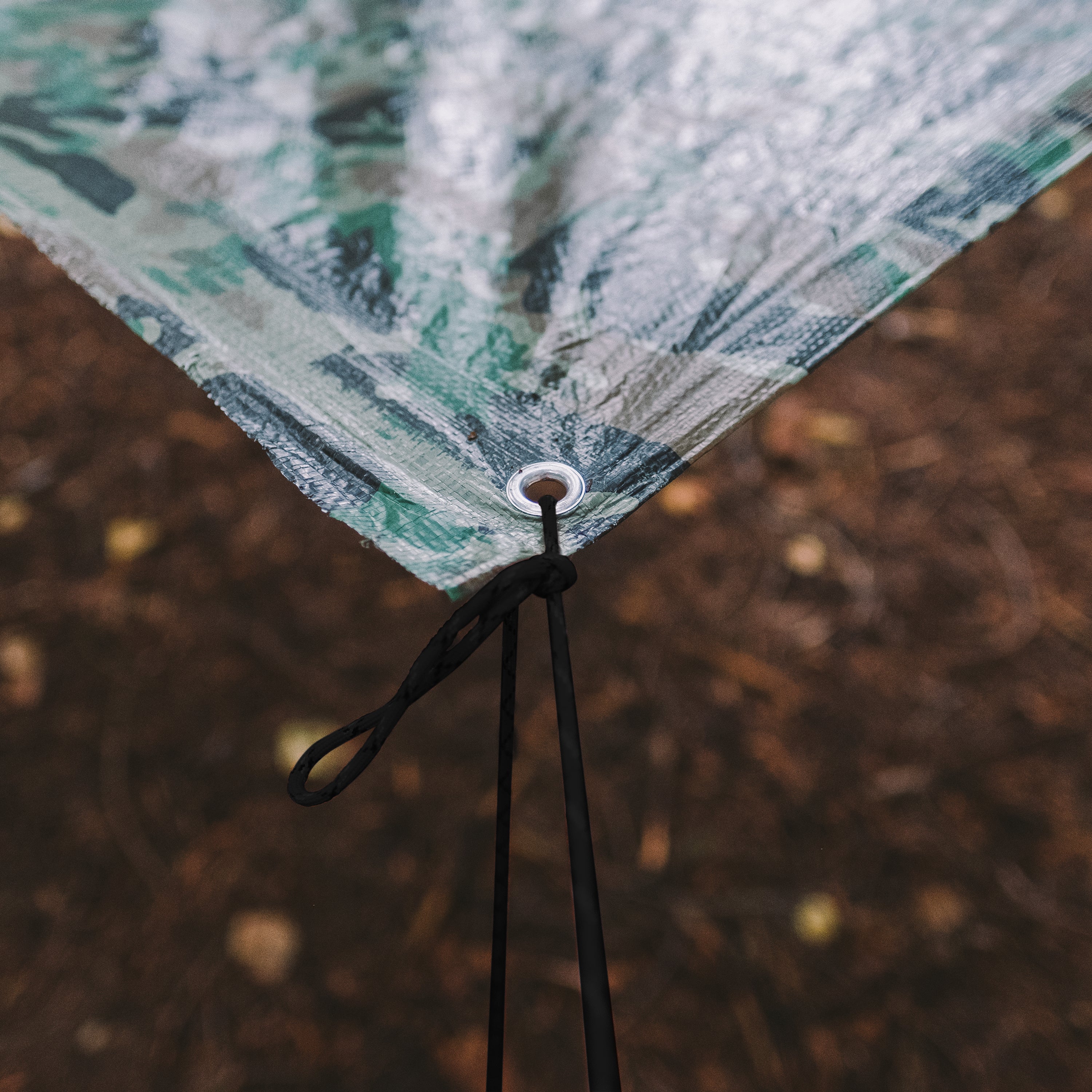 GearAid paracord tied to the corner of a camouflage tarp above a brown forest floor