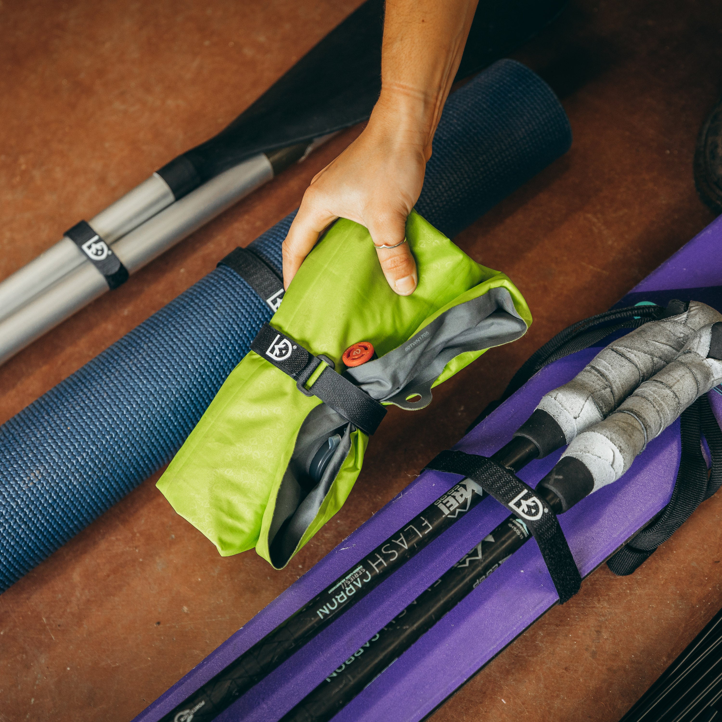 Hands placing an inflatable camp chair wrapped up with Fast Wrap on the ground inbetween other gear held in place with Fast Wraps like ski poles and skis, a yoga mat, and kayak paddles