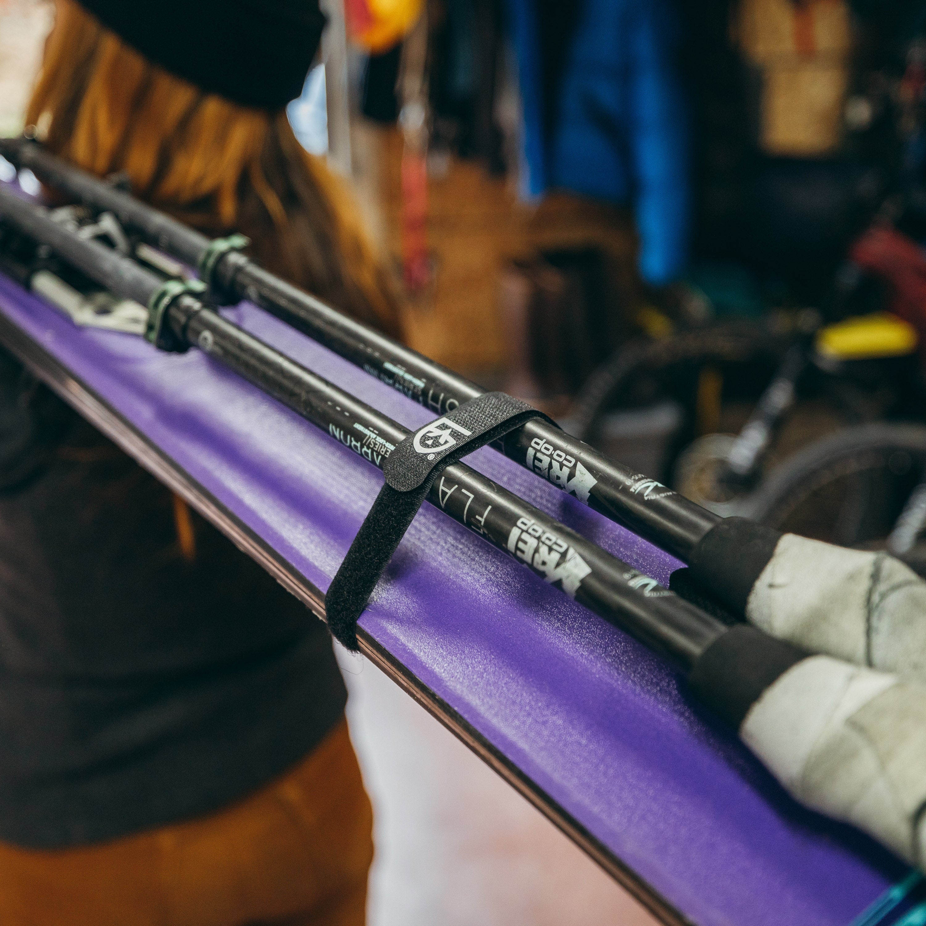 Person walking away with a pair of skis and ski poles strapped together with Fast Wrap, background is off a blurry gear garage