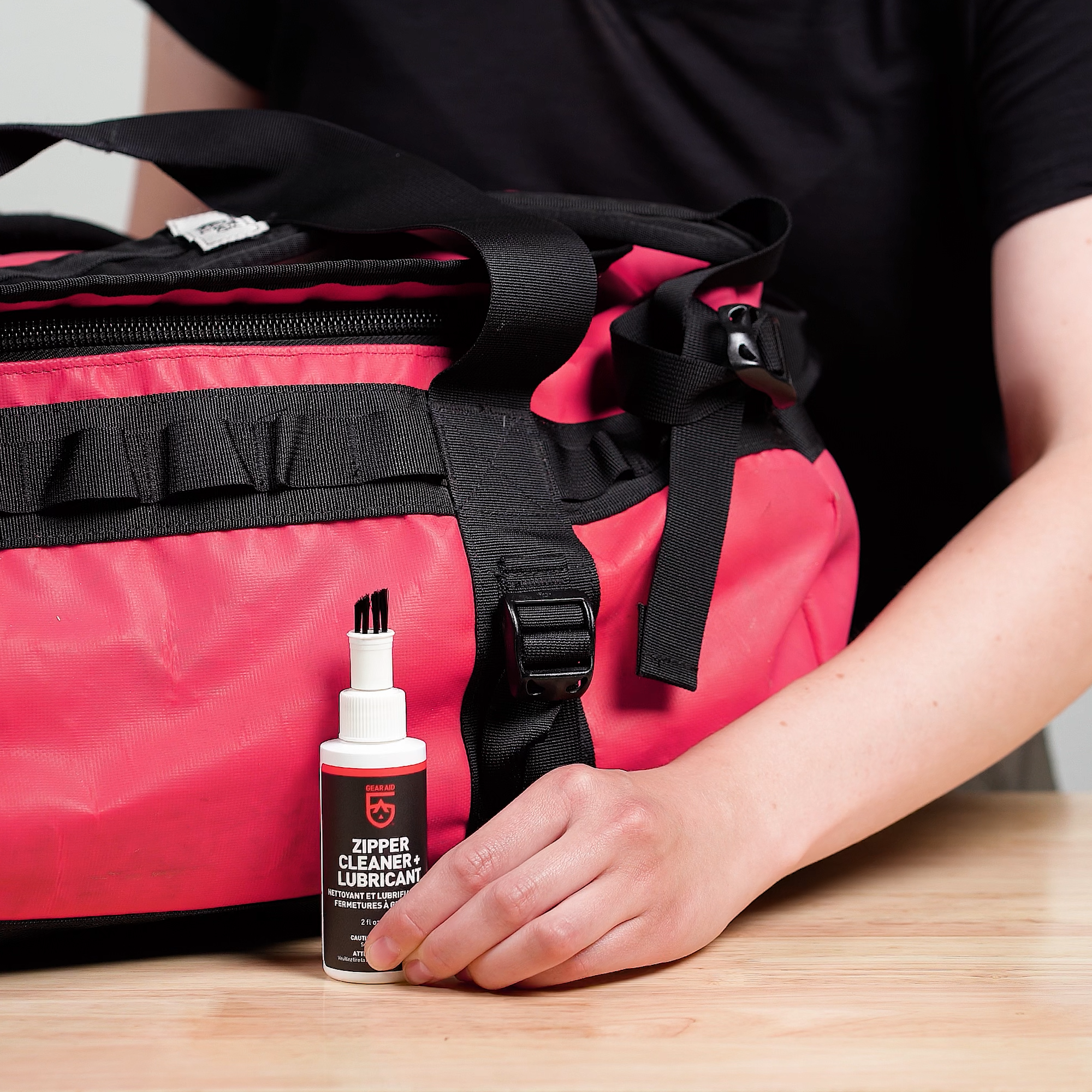 Hand holding bottle of GEAR AID Zipper Cleaner + Lubricant in front of red duffle bag