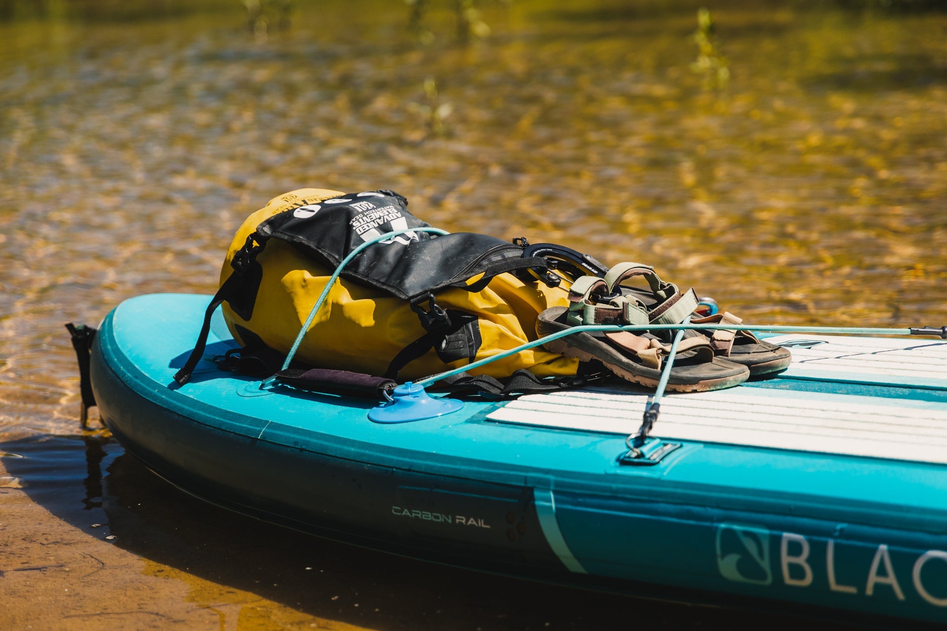 Blue paddle board in shallow water with a dry bag and sandals attached to it
