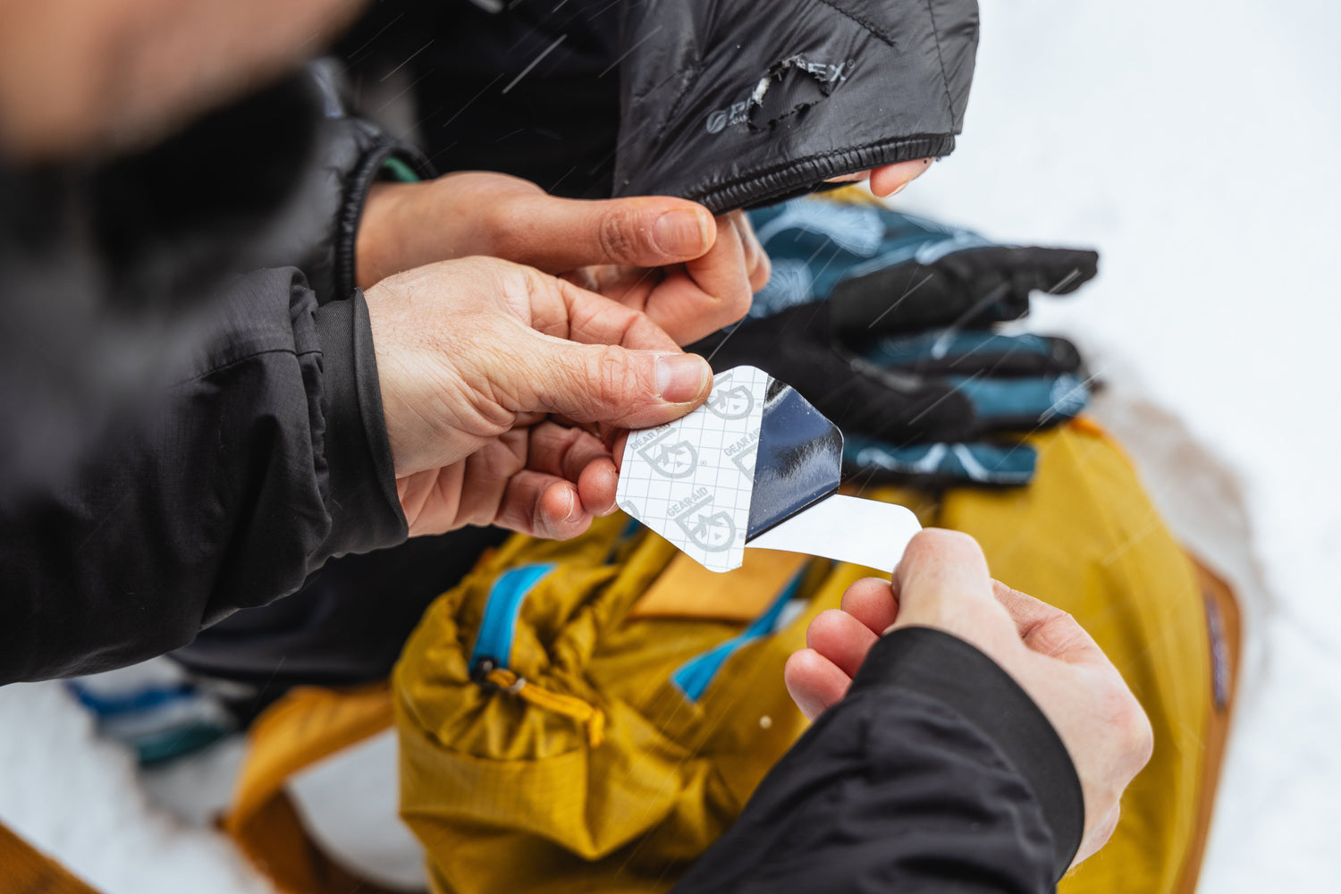 Image of a woman peeling a GORE-TEX fabric patch backing off a patch in the snow above a yellow pack