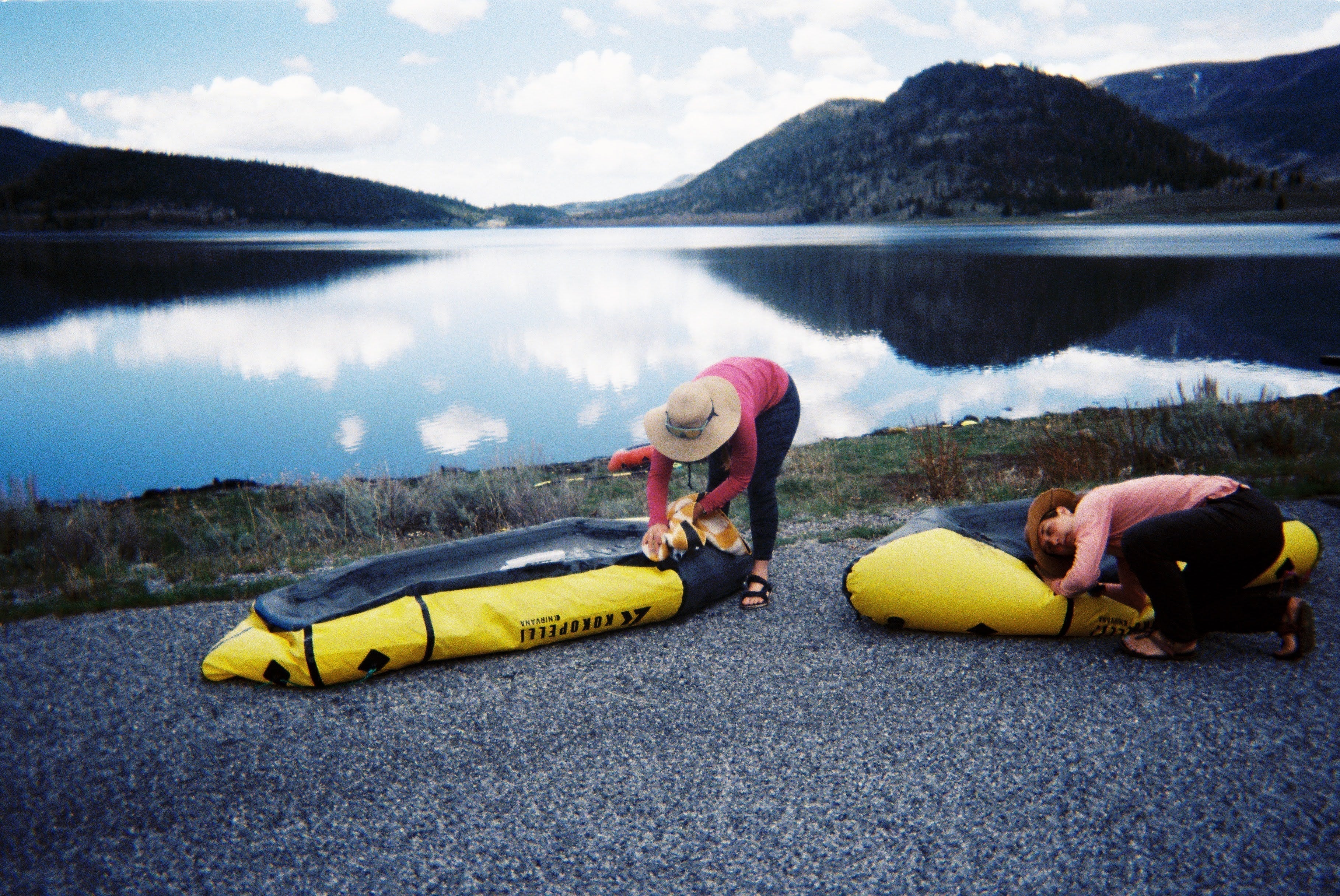 Two women standing on a gravel road before a lake blowing up Kokopelli Packrafts, on film