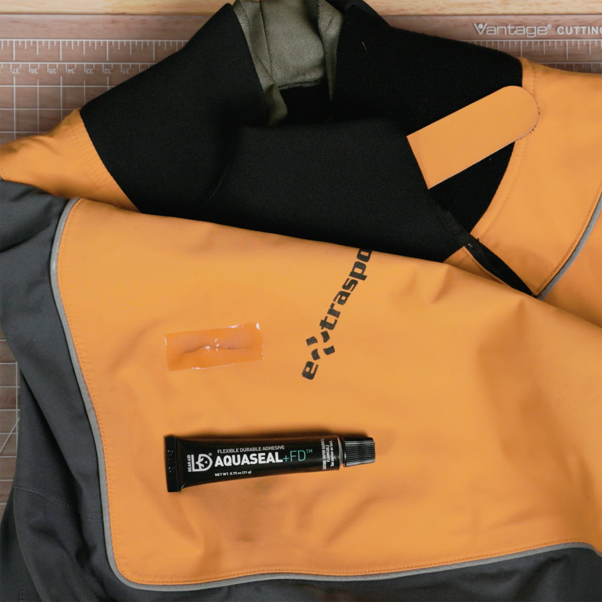 A small tube of Aquaseal FD sitting on a repaired orange, black, and gray drysuit top