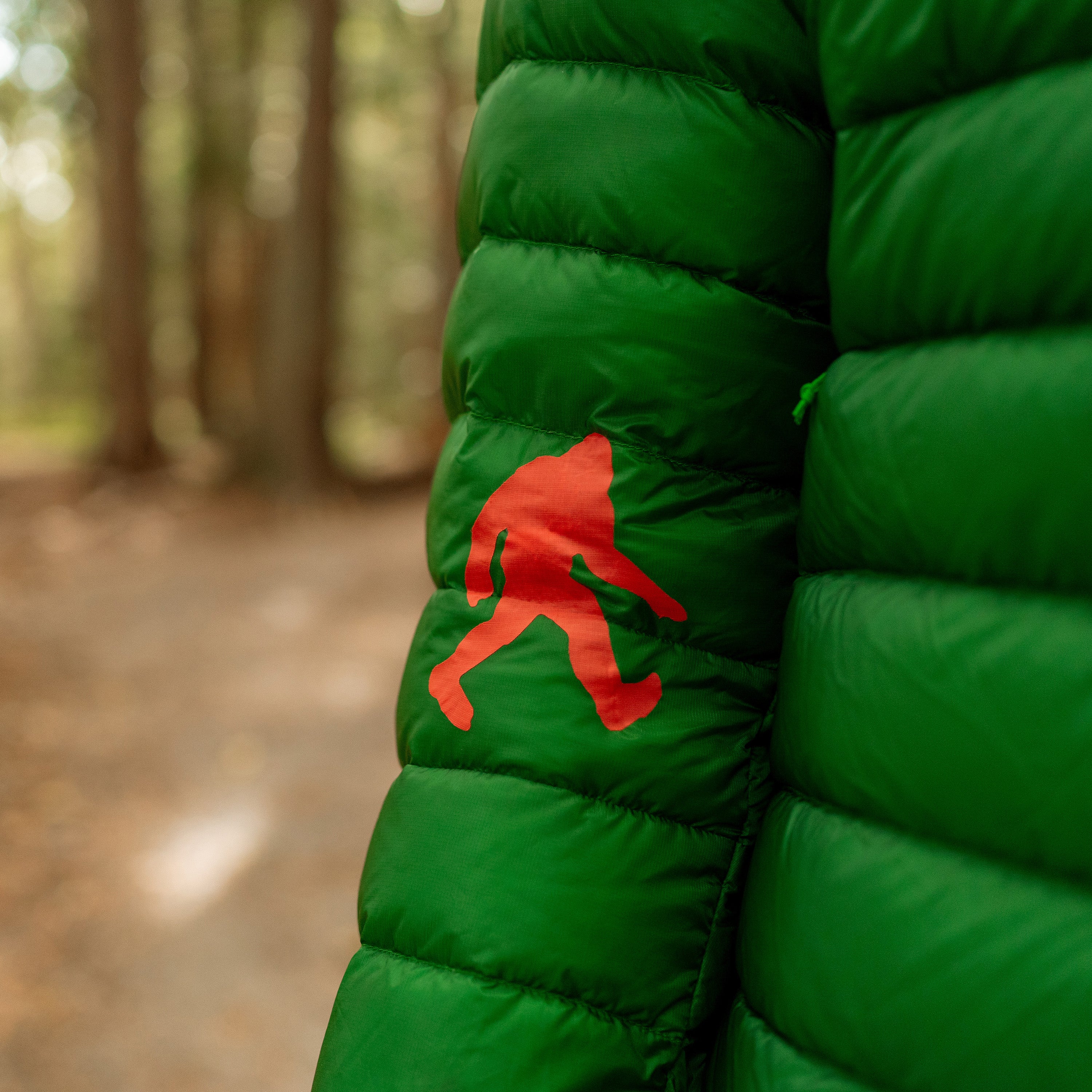 Green puffer jacket with a red sasquatch patch in a forest setting