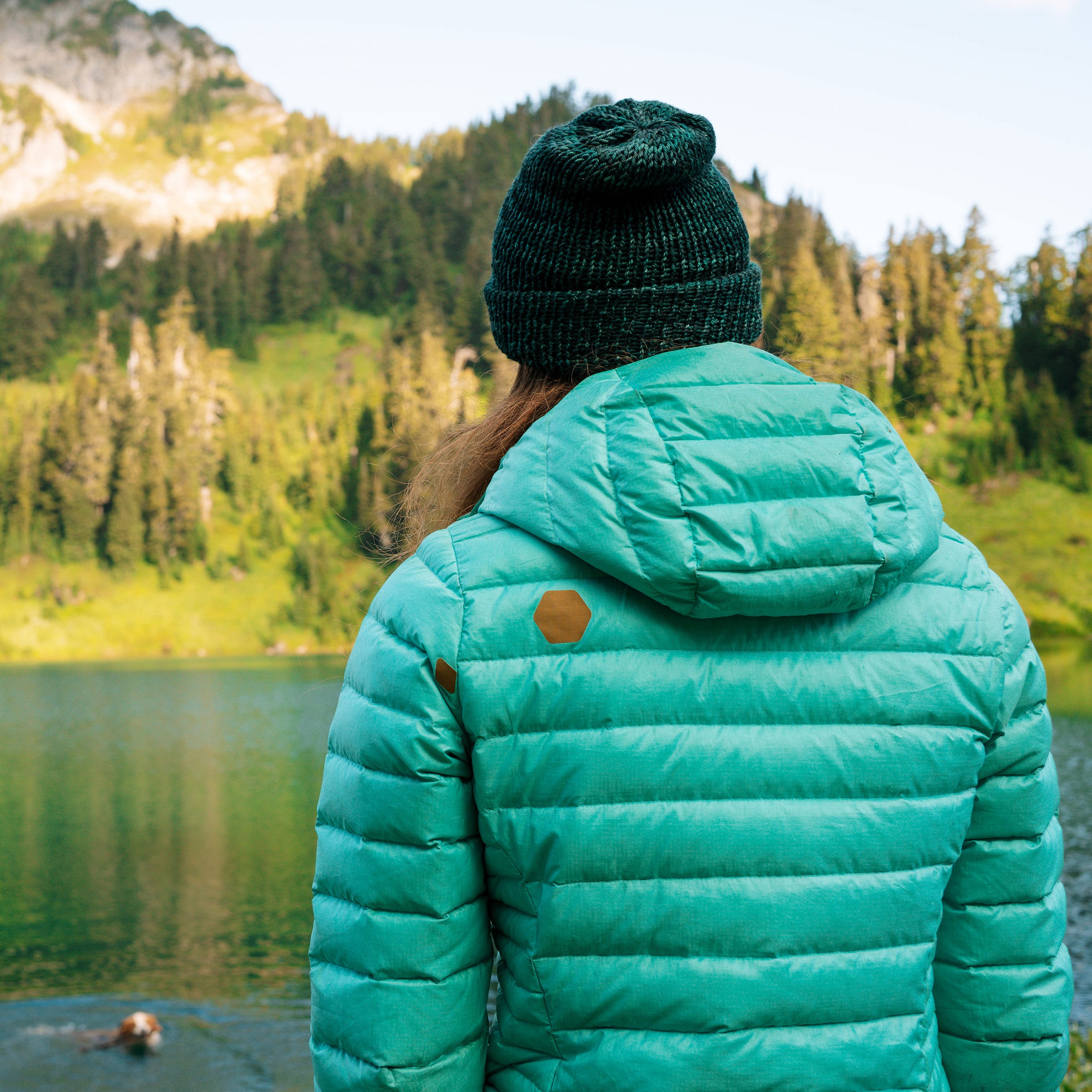 Person wearing a teal puffer jacket and green beanie standing by a lake with mountains in the background