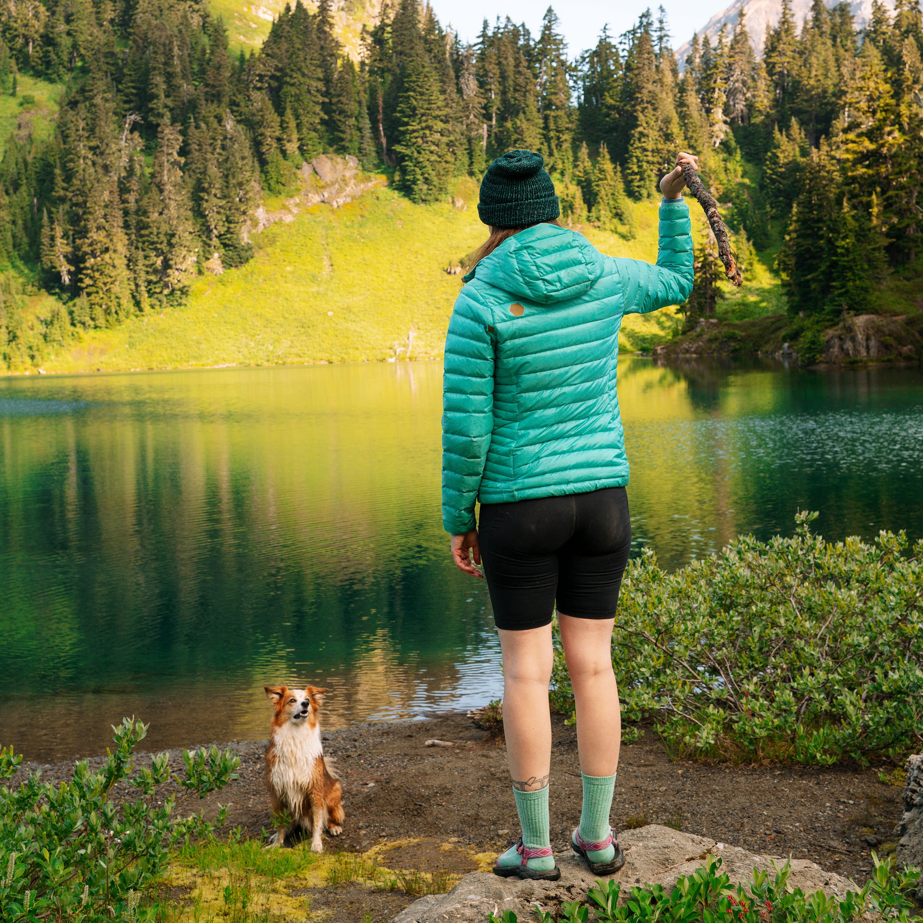 Person in a teal jacket standing by a lake with a dog, surrounded by trees and nature.