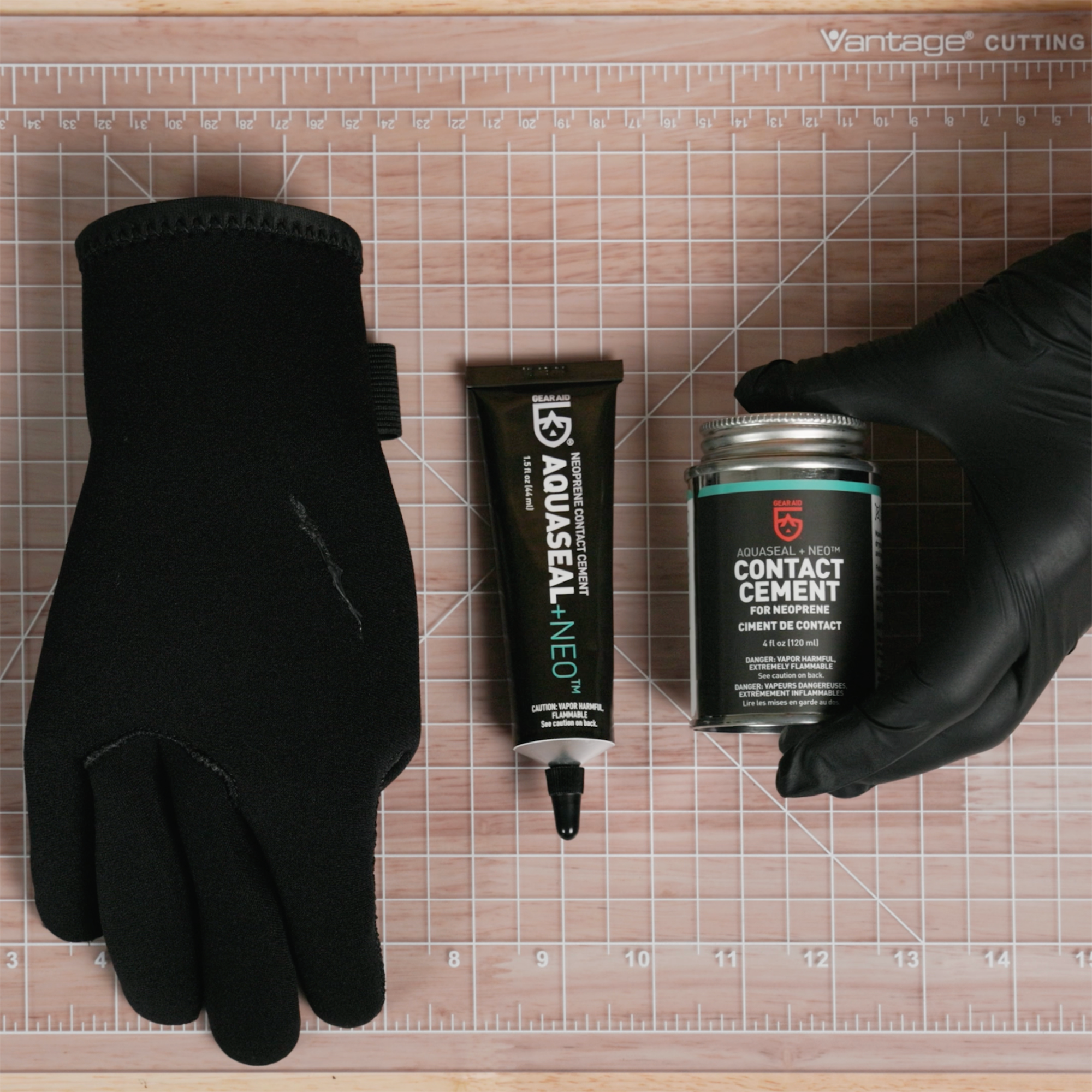Black neoprene glove with a repair on it, next to a tube of Aquaseal NEO and a tub of Aquaseal NEO being placed next to it by someone wearing a black glove