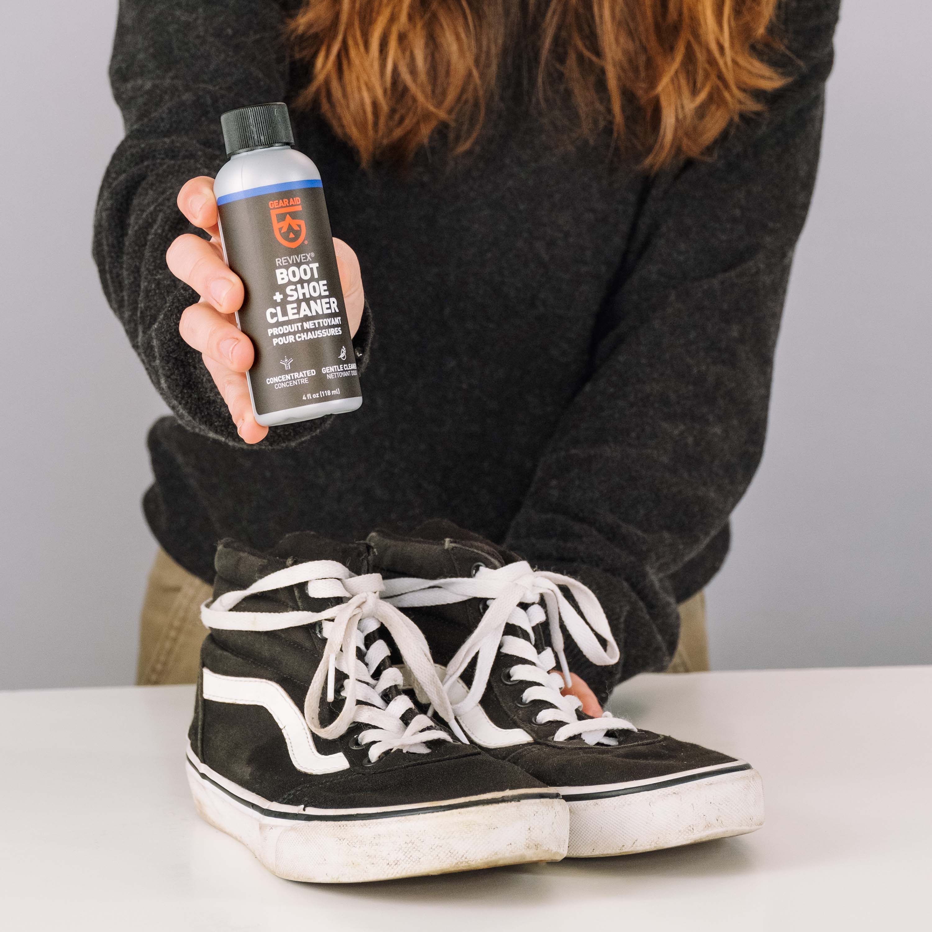 Person holding a bottle of boot and shoe cleaner next to a pair of black high-top sneakers on a table.