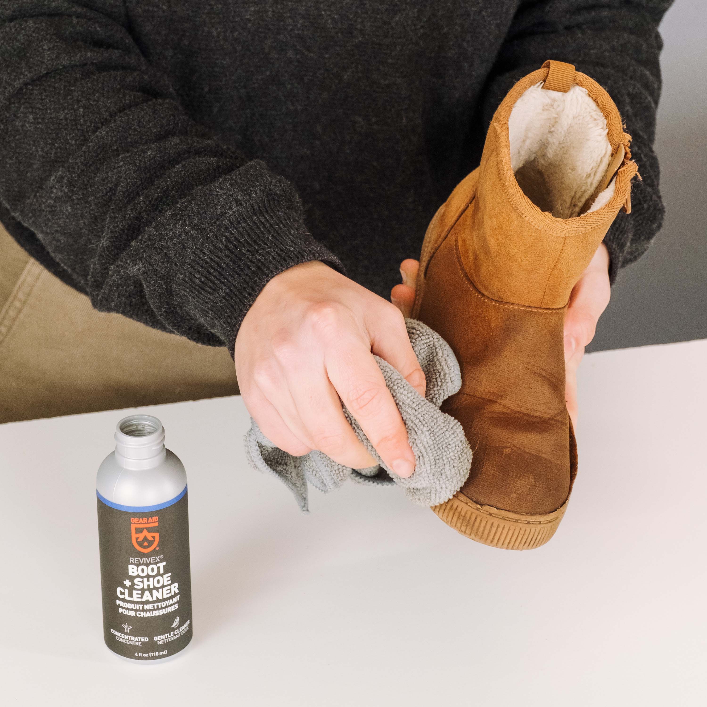 Person cleaning a brown boot with a rag, next to a bottle of boot cleaner on a white surface.