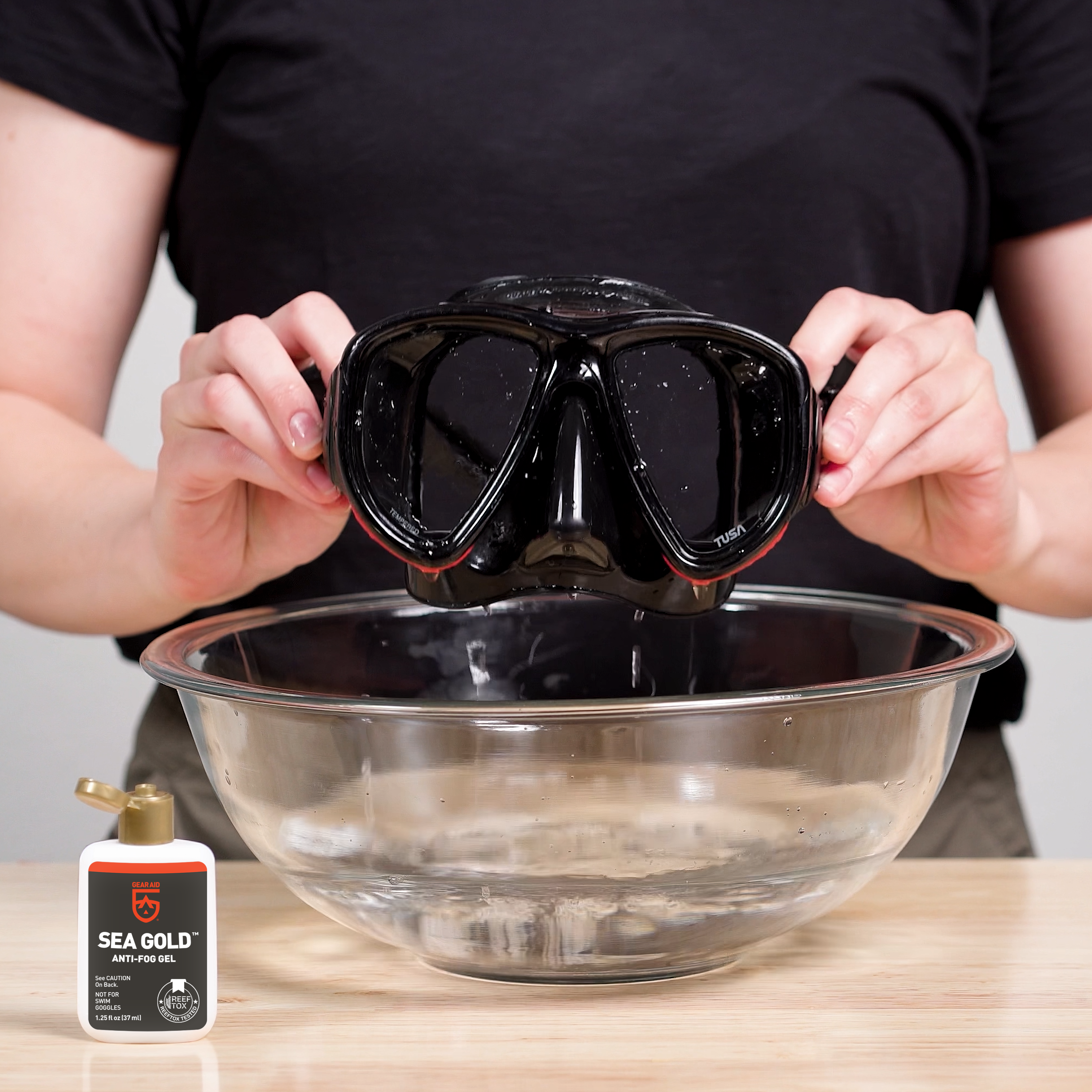 Dive mask after treatment with GEAR AID Sea Gold Anti-Fog Gel, held above a bowl of water next to the product bottle
