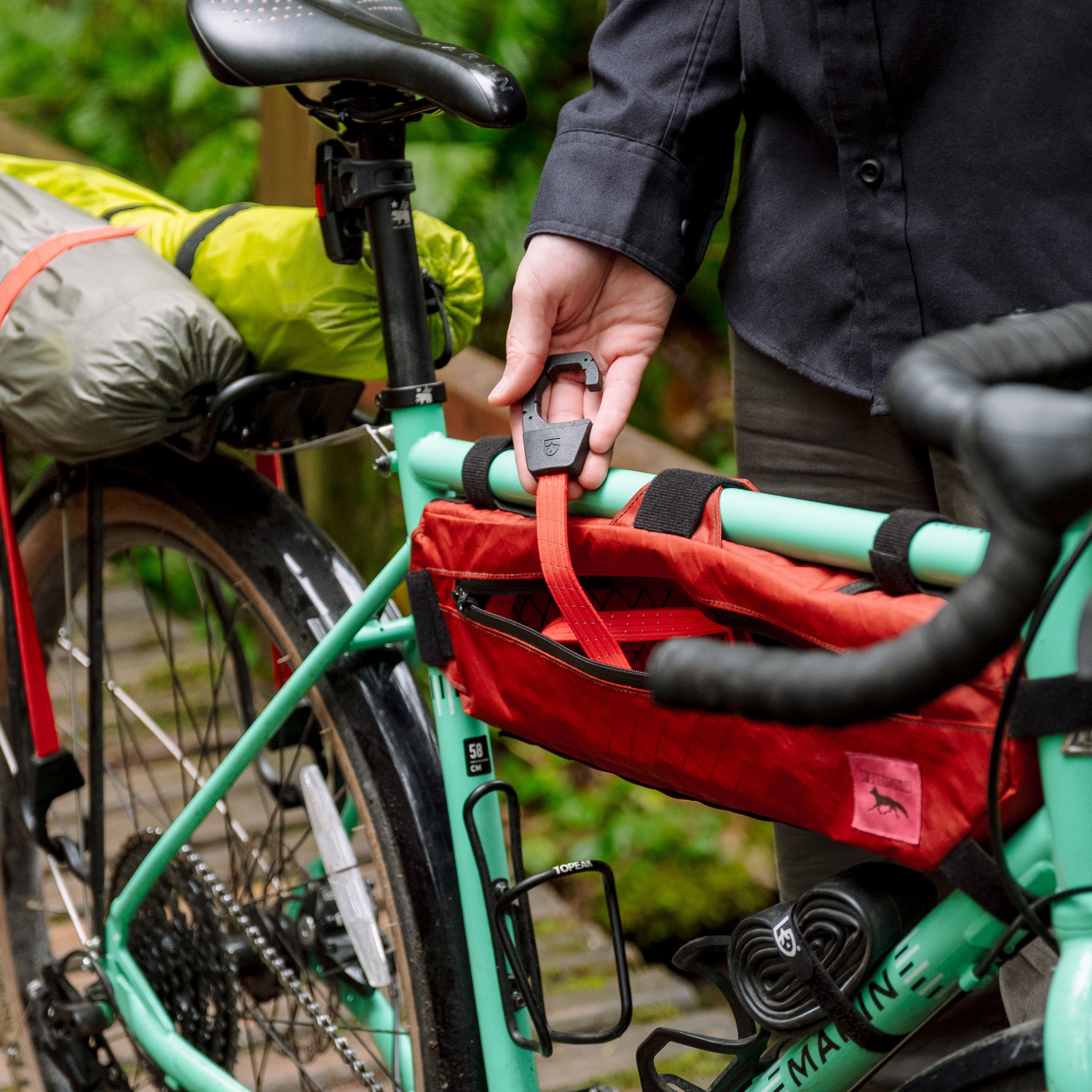 Person pulling a holdfast strap out of a red bike frame bag on a teal Marin gravel bike while standing on a bridge
