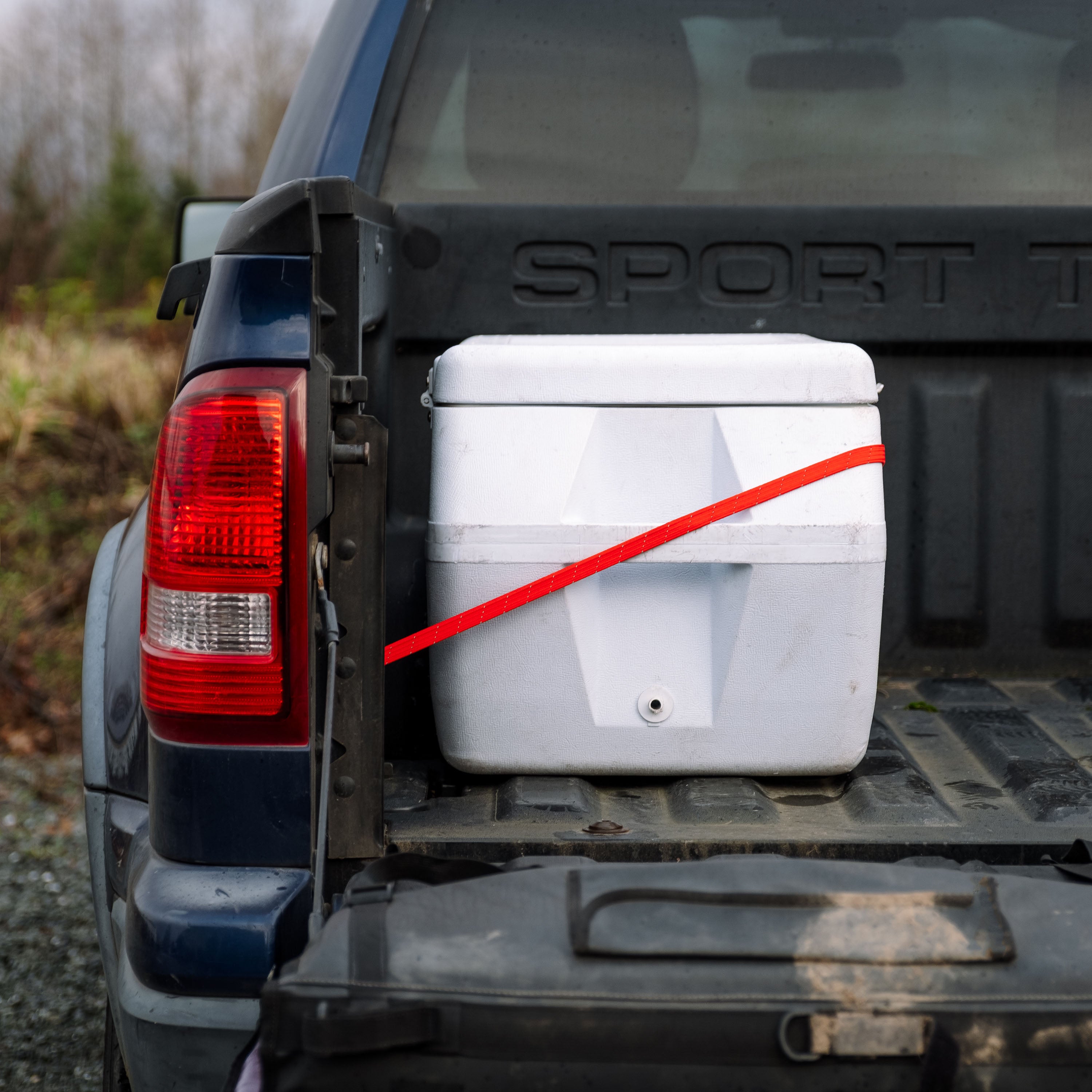 Cooler in the back of a blue pickup truck being held in place with a red Holdfast Strap, fall foliage in the background