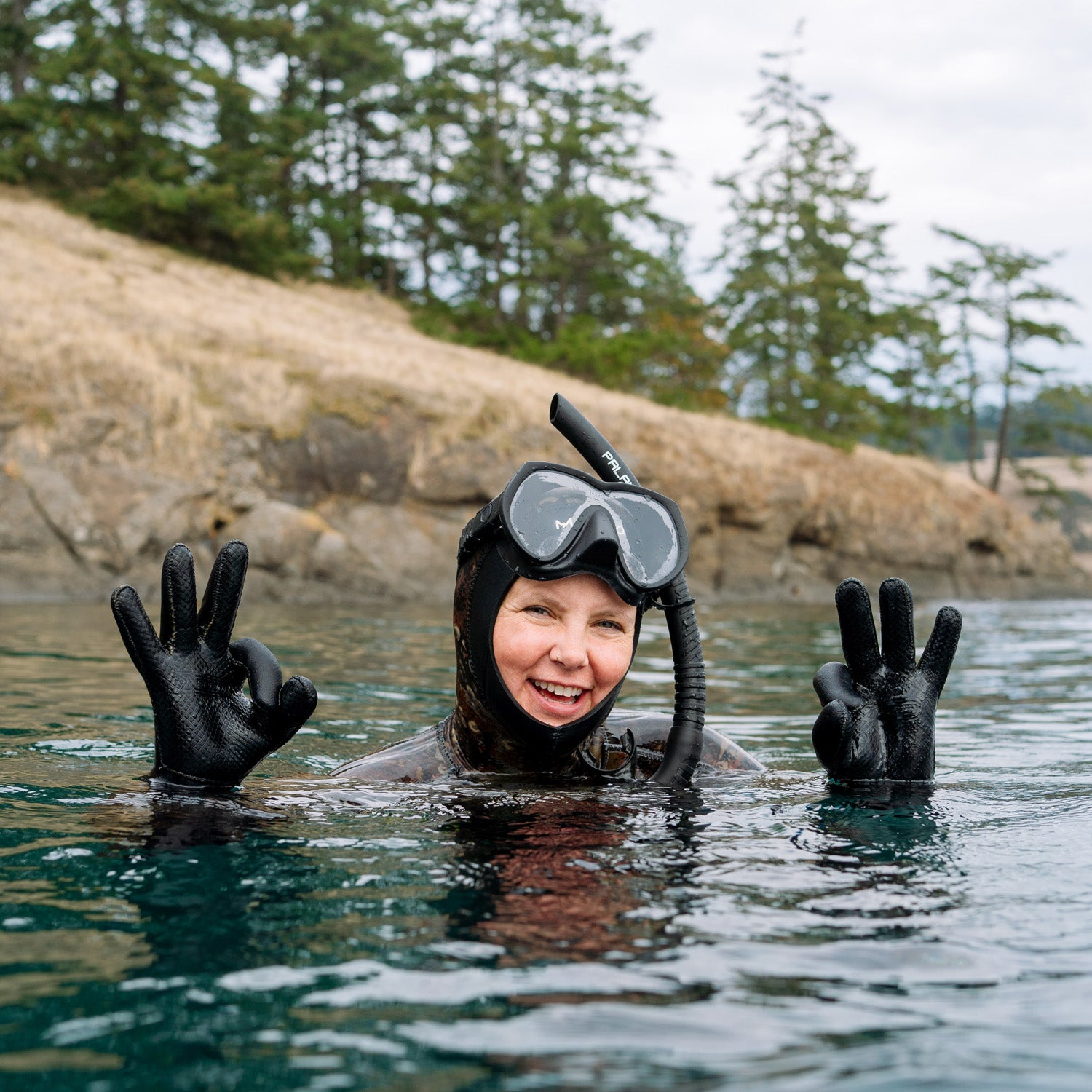 Person in a wetsuit and snorkeling gear waving from the water with a scenic background.