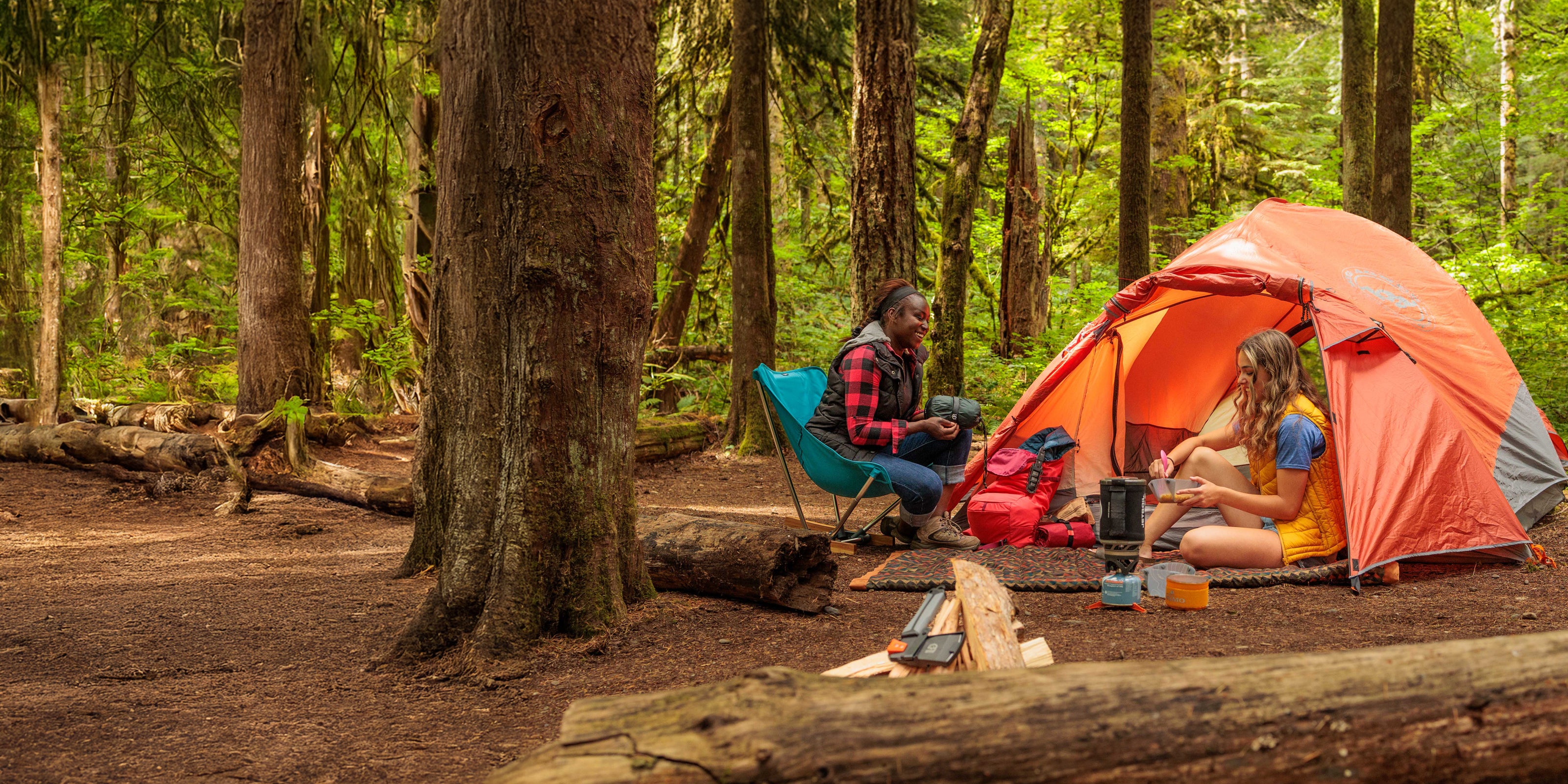 Two people camping in a forest with an orange tent and chairs.