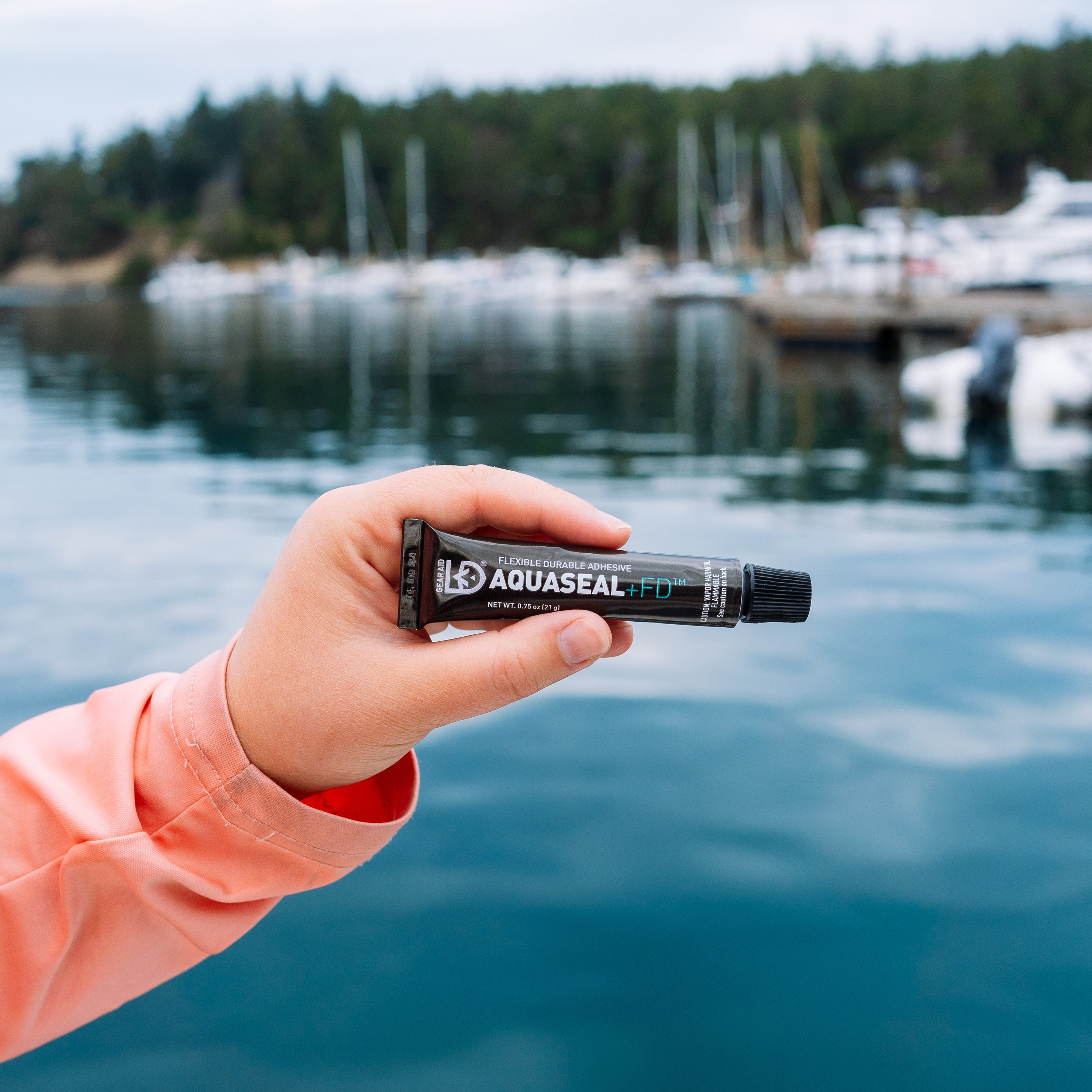 Hand holding a tube of Aquaseal by a marina with boats and water in the background