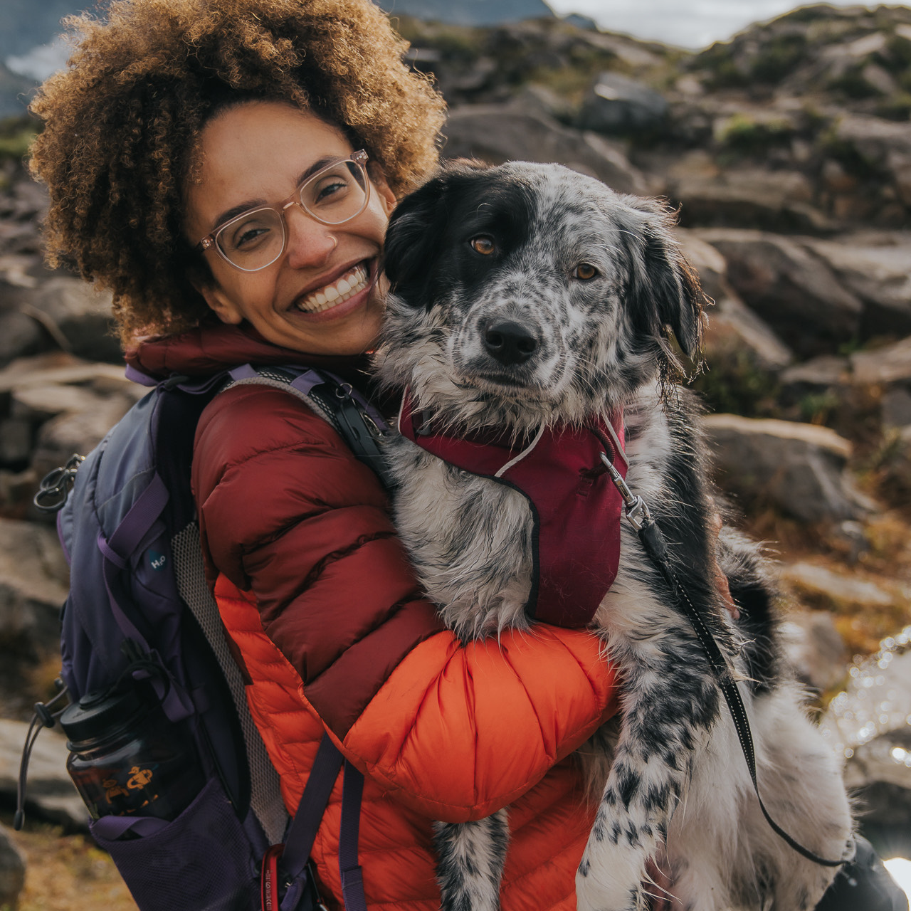 Woman in a red jacket holding an adorable dog in a mountainous landscape.
