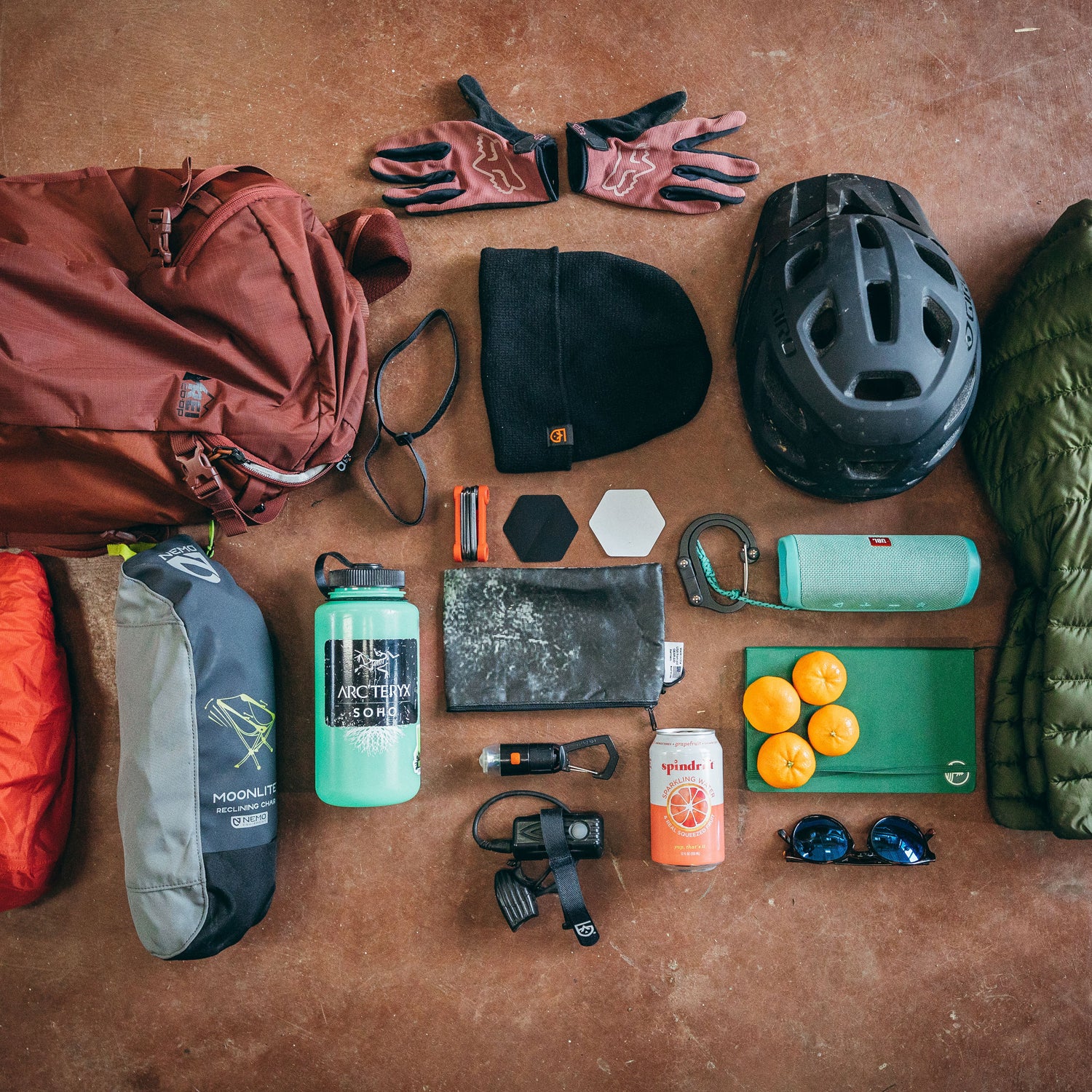 Collection of outdoor gear including a backpack, gloves, helmet, water bottle, and snacks on a brown surface.