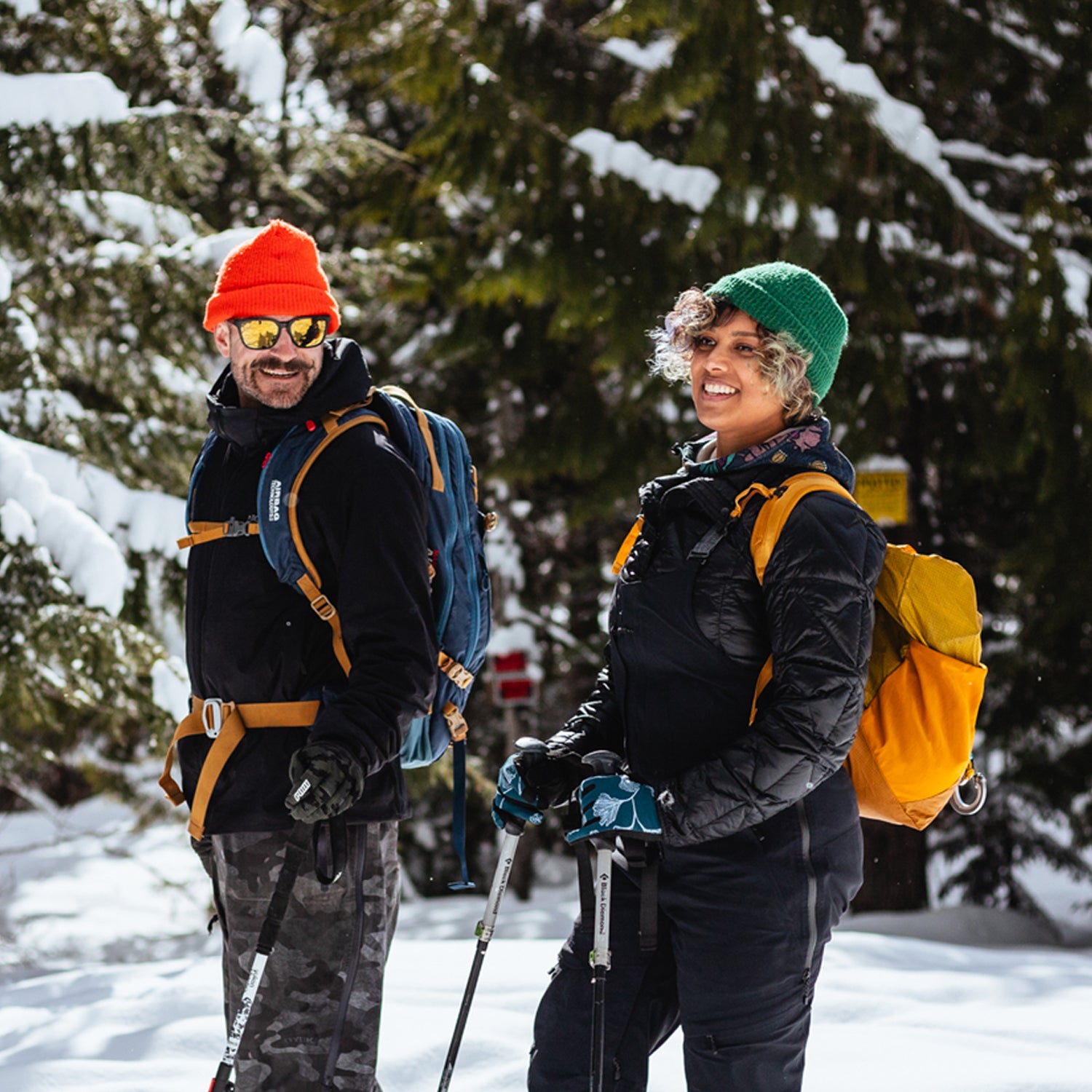 Two people with backpacks and ski poles standing in a snowy forest.
