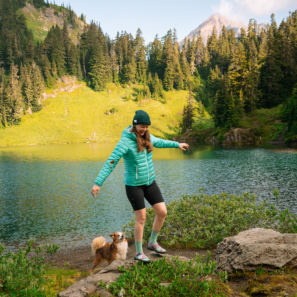Person in a green jacket standing next to a dog by a lake with mountains in the background.