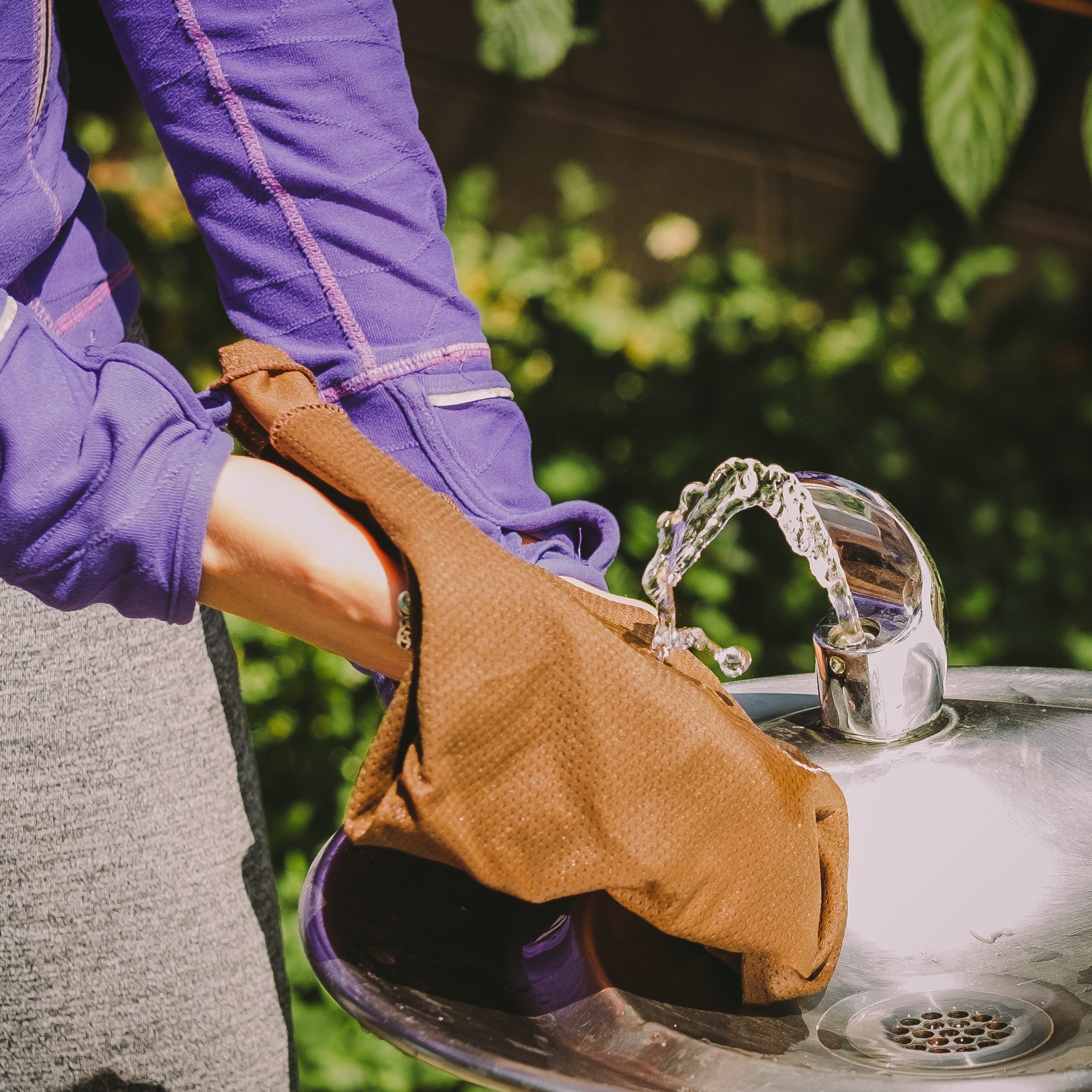 Person washing a brown leather bag over a sink with water flowing.