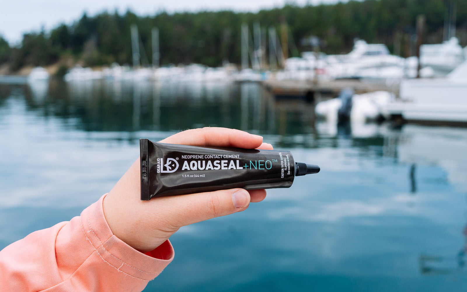 Person holding a tube of Aquaseal Neo by a marina with boats in the background