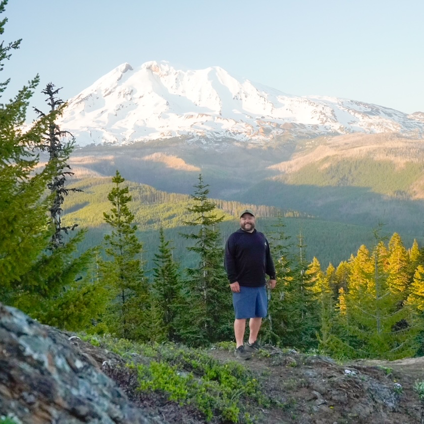 Person standing on a mountain trail with snow-capped mountains in the background.