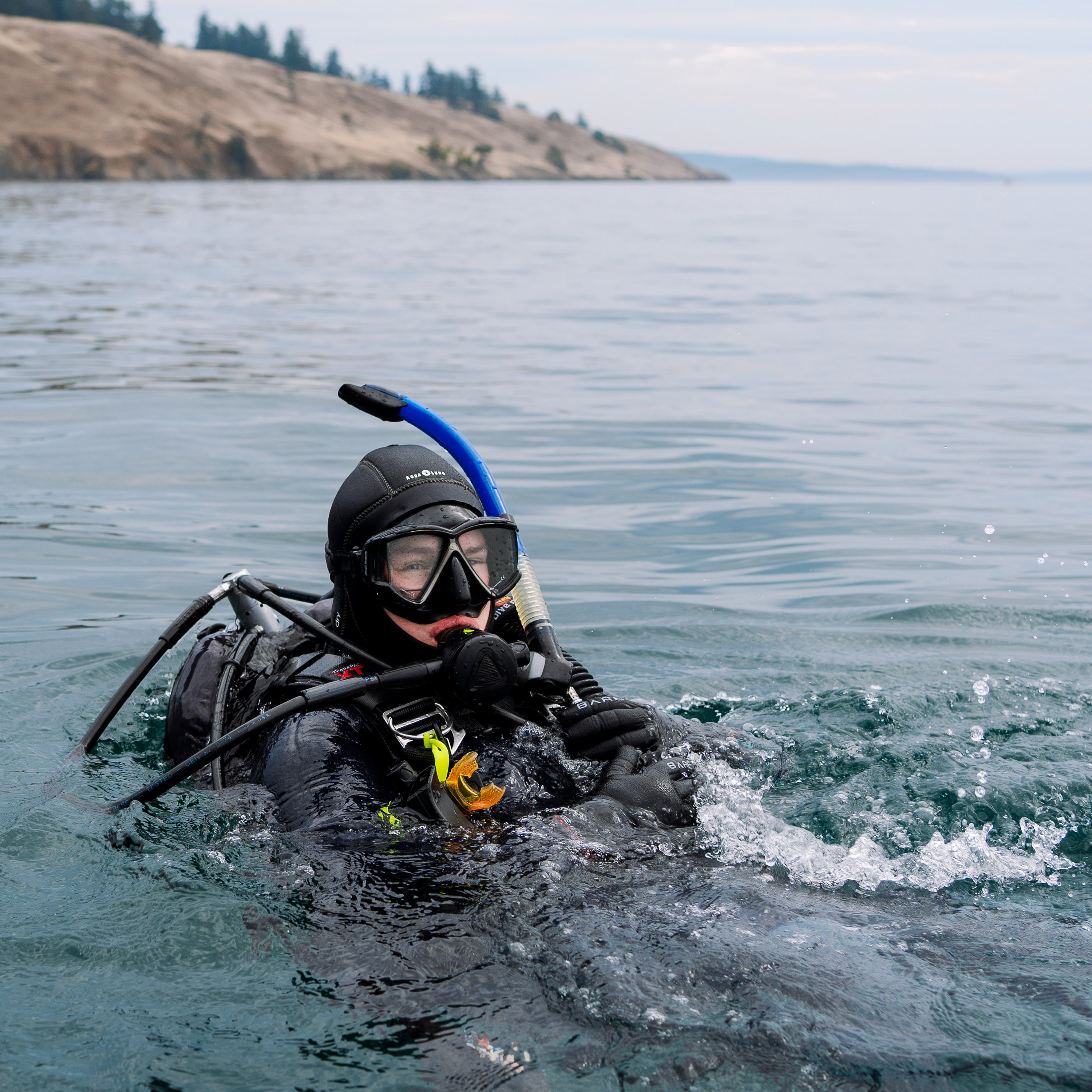 Person in scuba diving gear emerging from water with a scenic background.