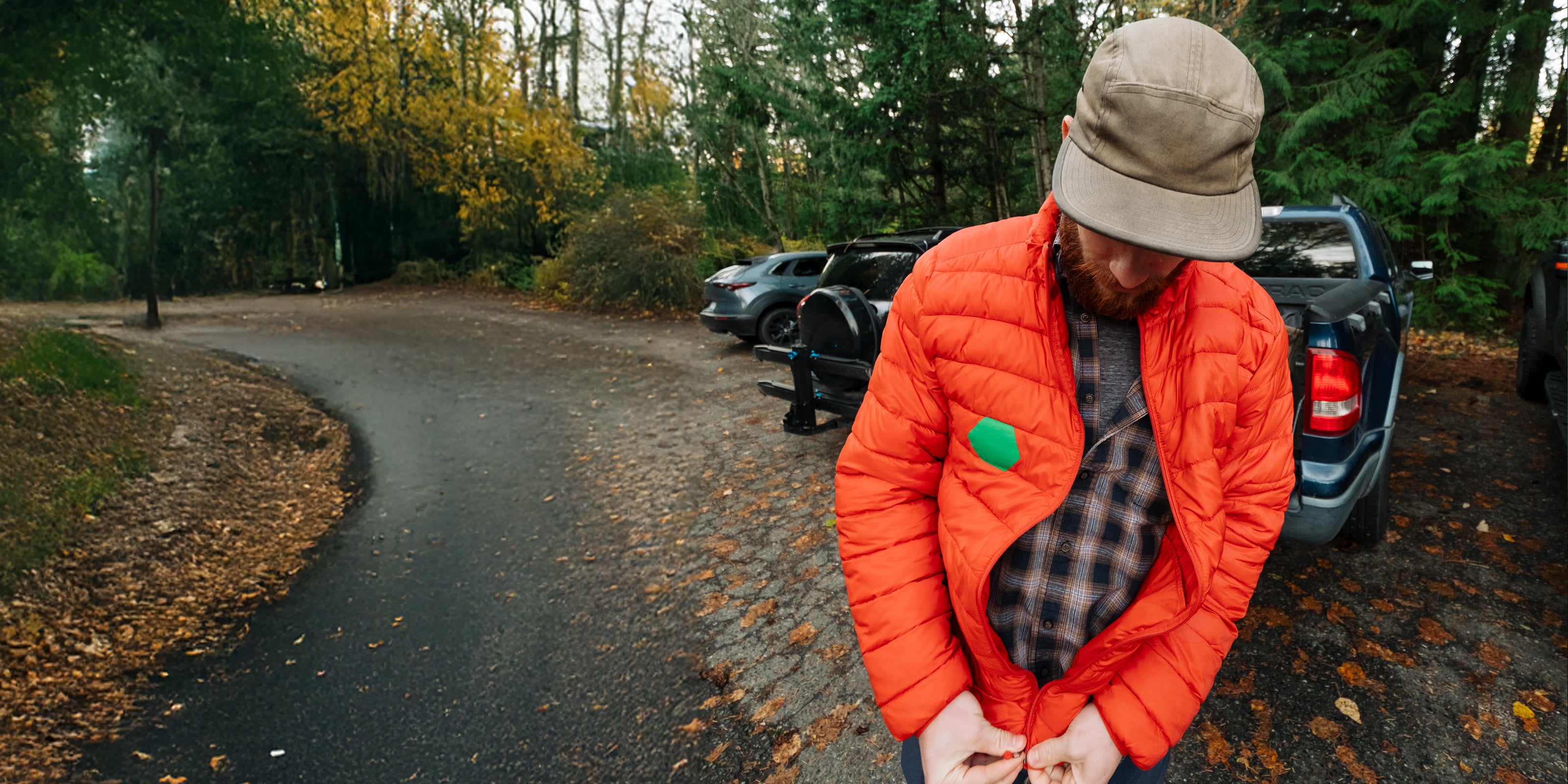 Person in an orange jacket and beige hat with green Tenacious Tape patch standing on a road with trees and parked cars in the background.