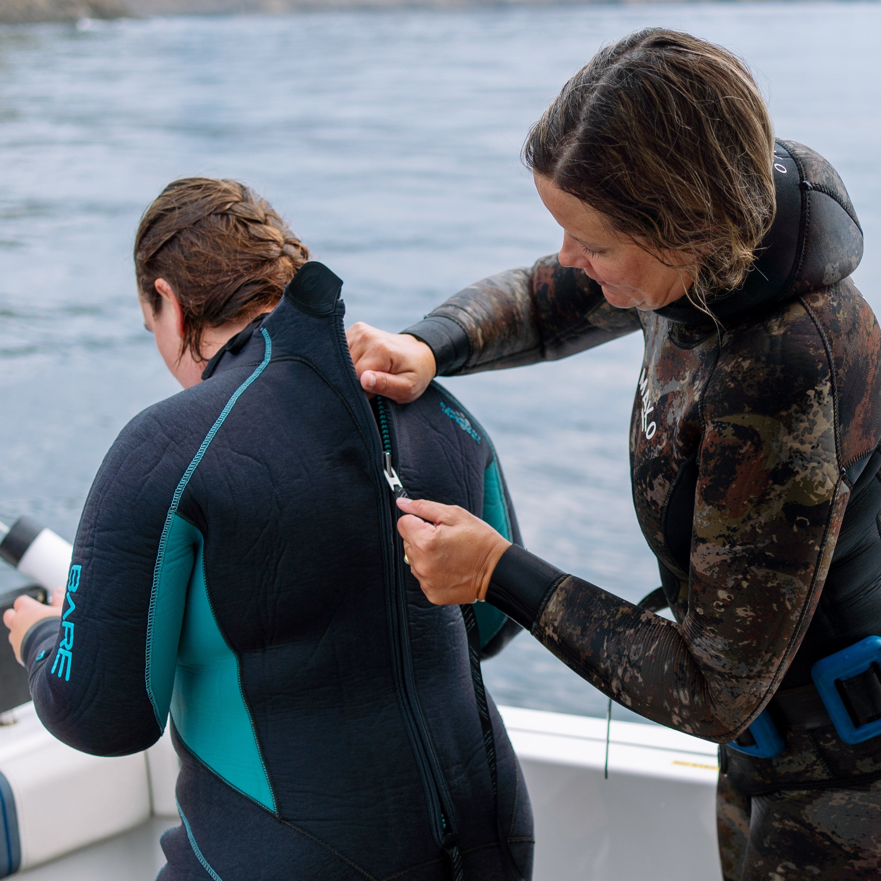 Person helping another person put on a wetsuit by a body of water.
