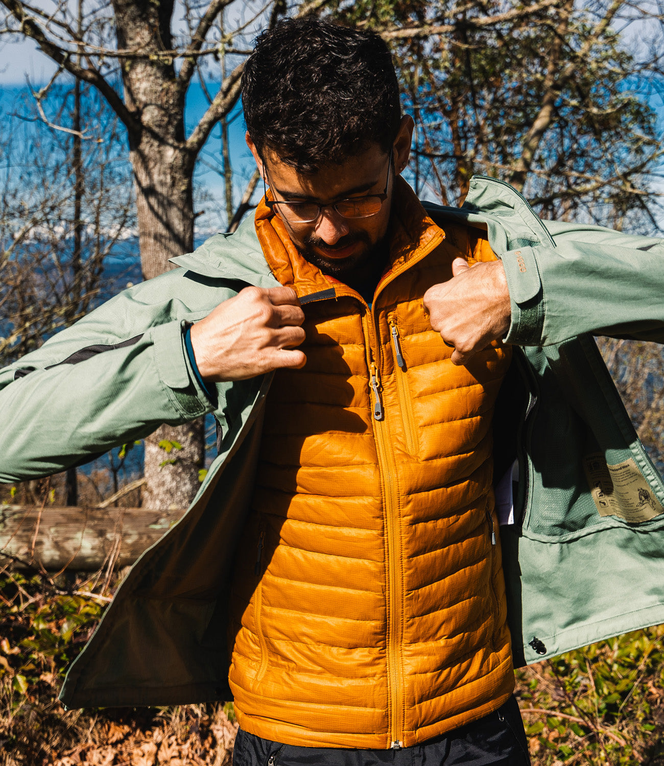 Person wearing a green jacket over an orange puffer vest outdoors with trees and water in the background