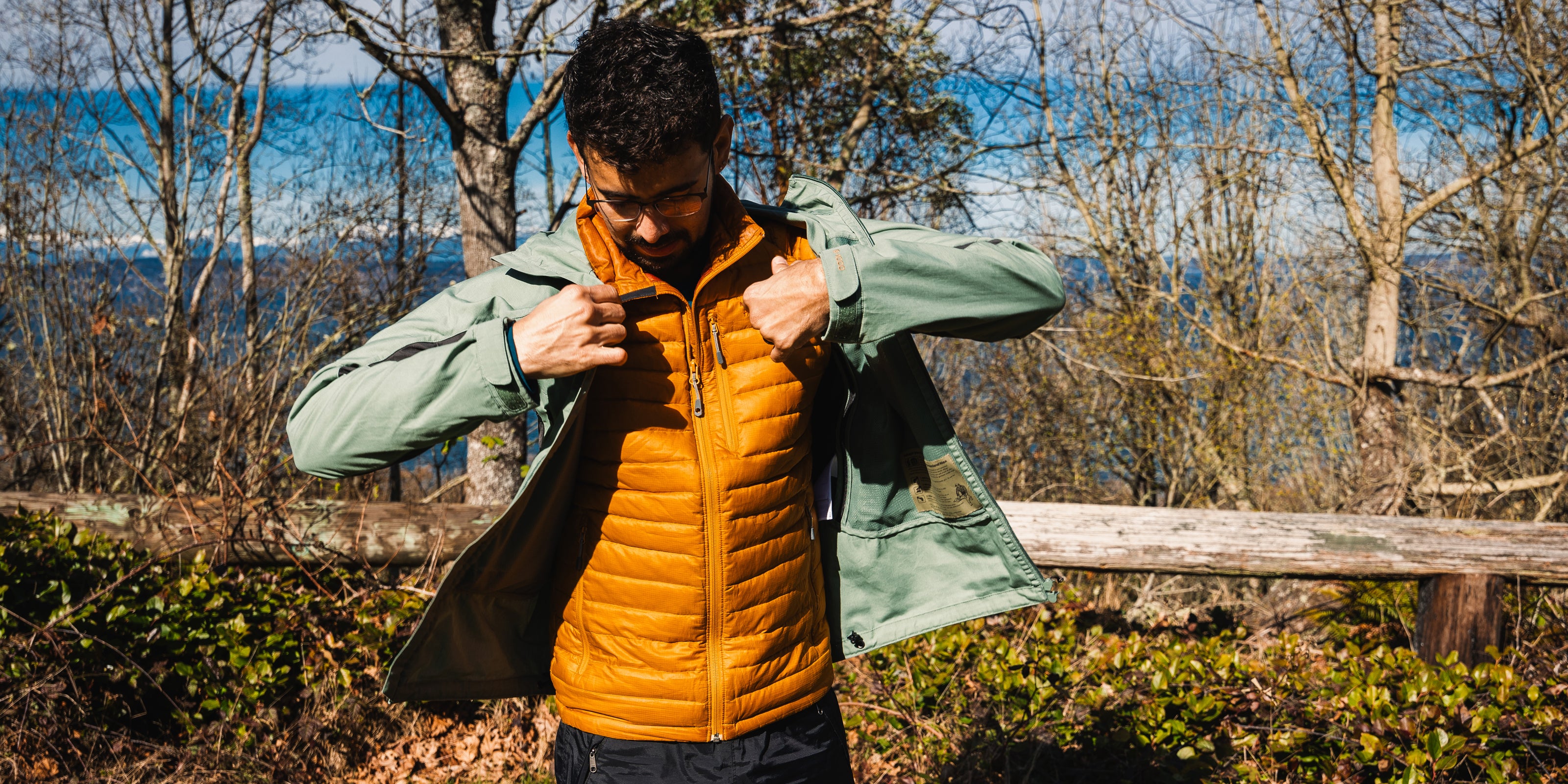 Person wearing a green jacket over an orange puffer vest outdoors with trees and water in the background.