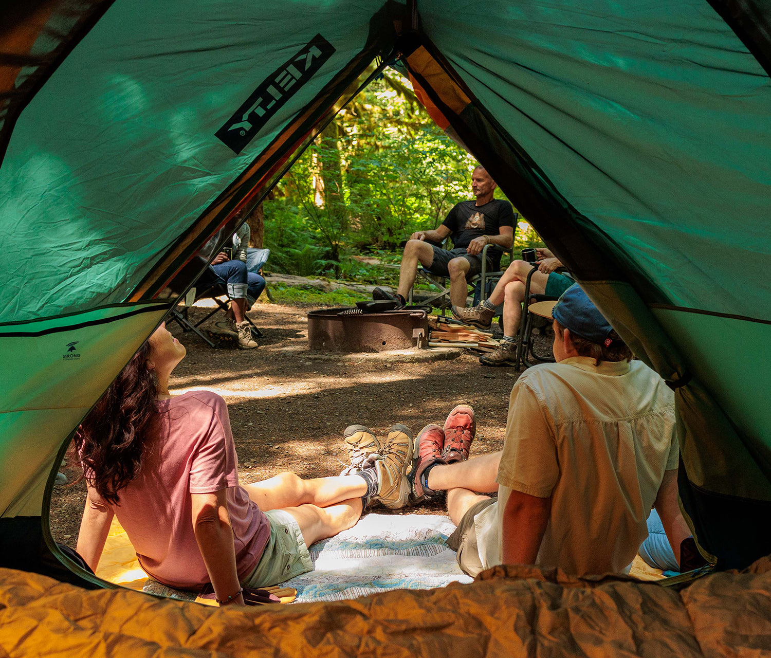 People camping outdoors, sitting around a campfire with a tent open.